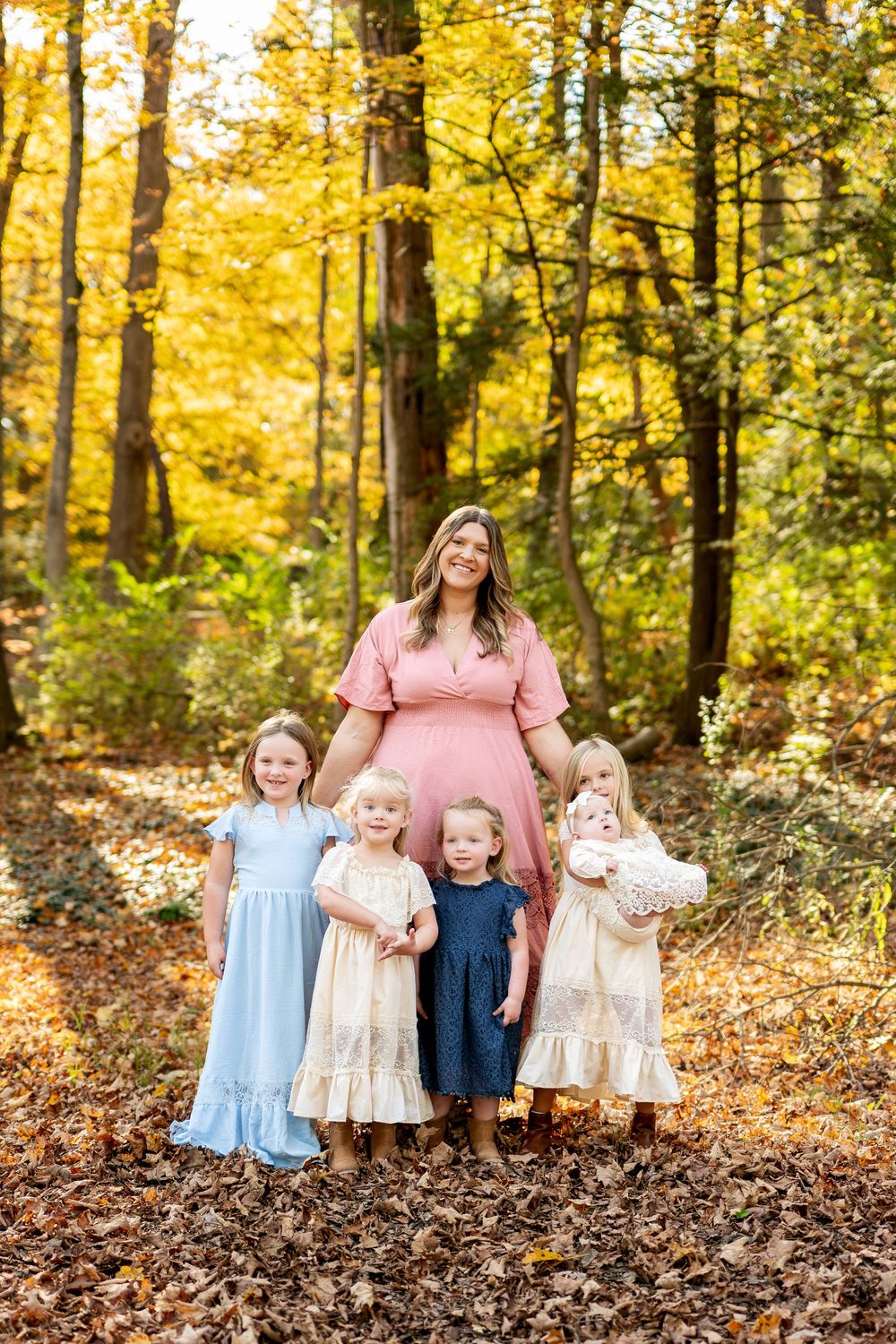 Mother in pink dress posing with four young daughters in an autumn forest setting.