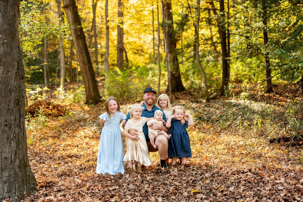 Father crouching with five young daughters among fall leaves in a sunlit autumn forest.
