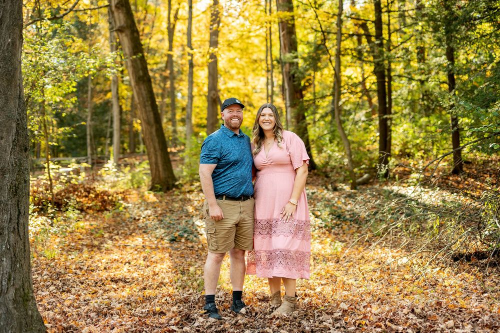 Couple smiling together in a sunlit autumn forest, man in blue shirt and woman in pink dress.