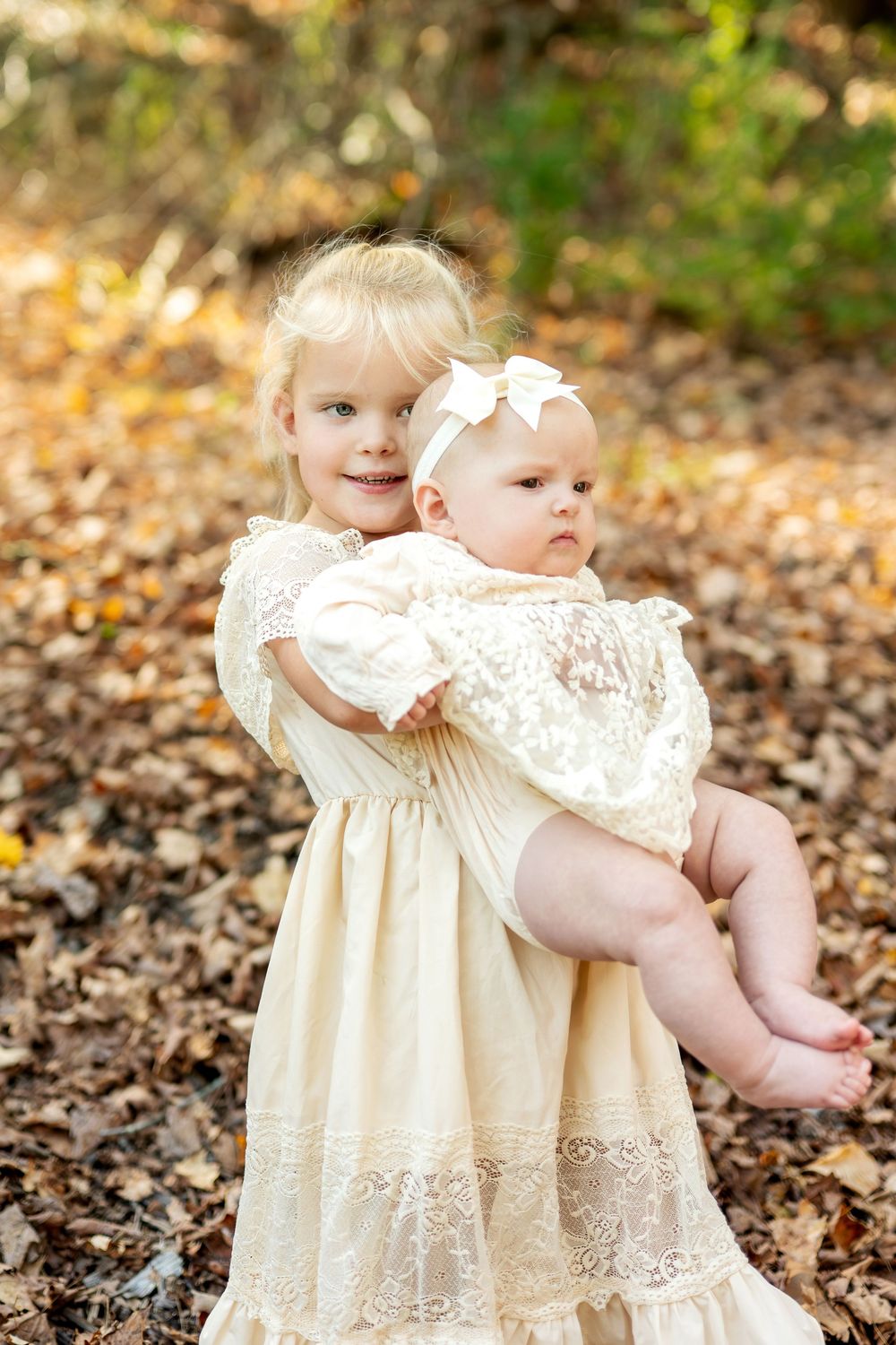 Young blonde girl in cream lace dress holding baby sibling in autumn forest.