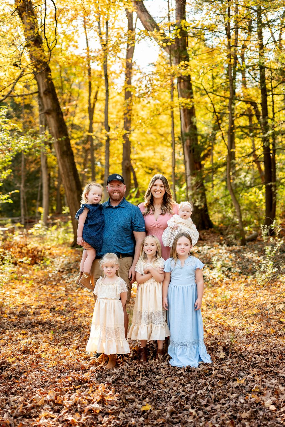 A family of seven poses among vibrant golden autumn trees with four girls and a baby in the woods.