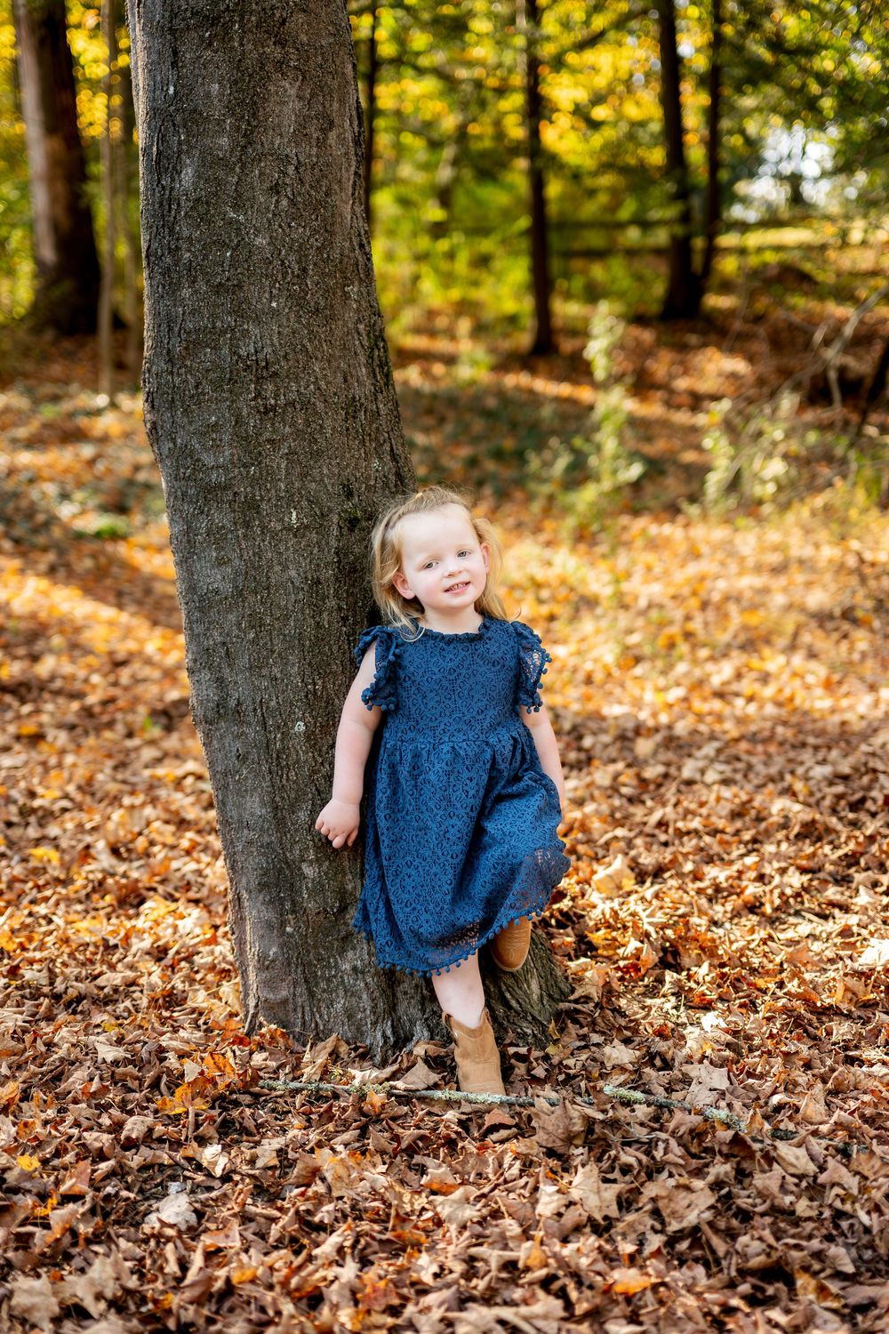 Toddler girl in navy blue dress standing barefoot against a tree in a fall leaf-covered forest.