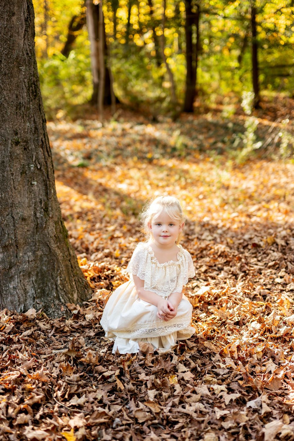 Young blonde girl in cream lace dress crouching among fallen autumn leaves in a sunlit forest.