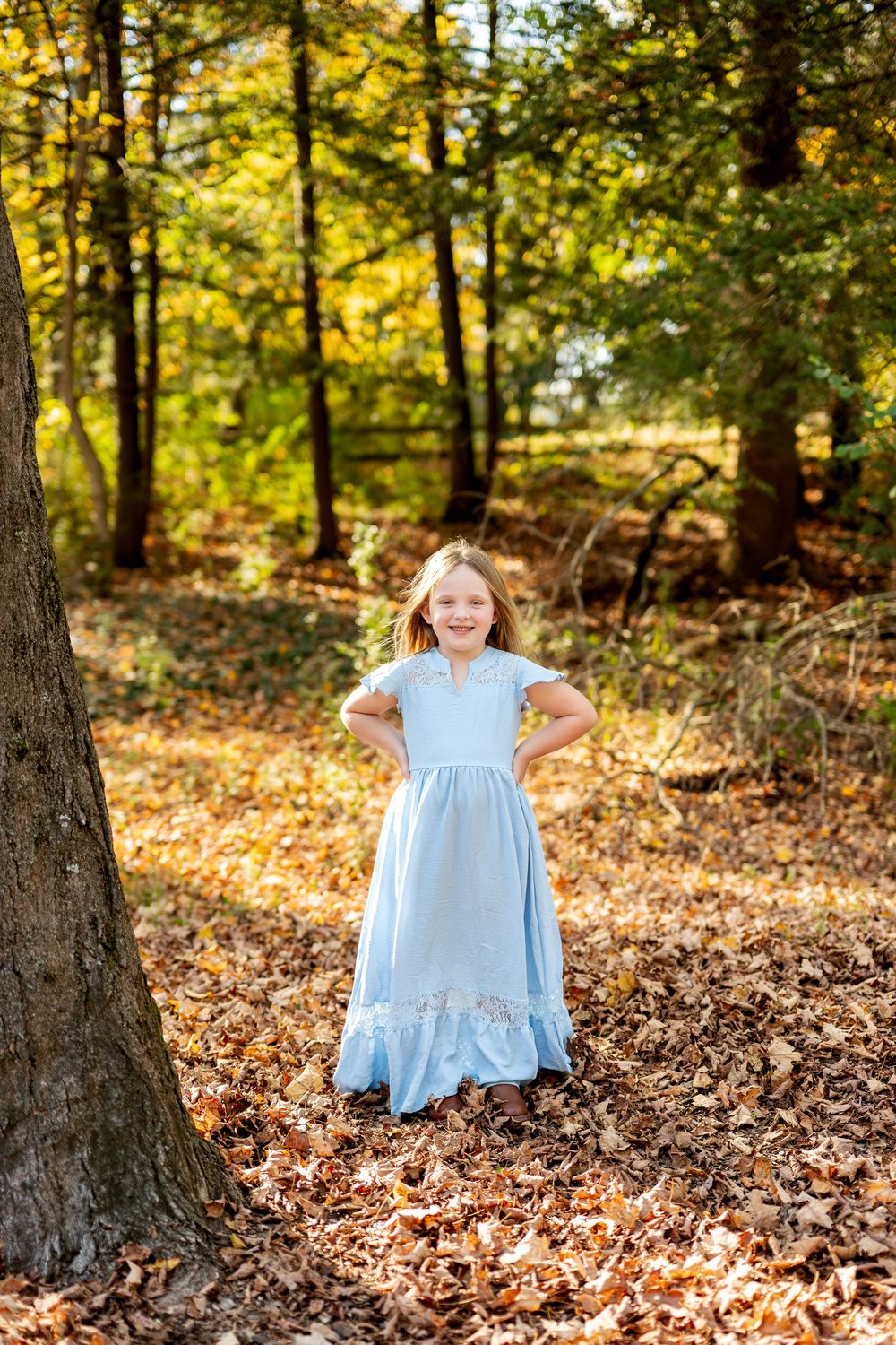 Young girl in light blue maxi dress posing with hands on hips among autumn leaves in a forest.
