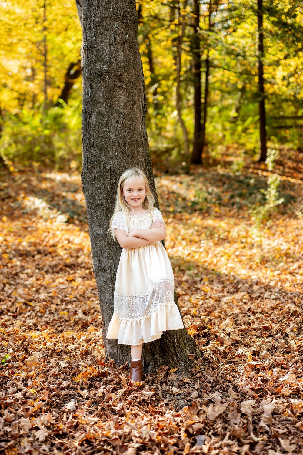 Blonde girl in cream dress leaning against a tree trunk surrounded by golden fall foliage.