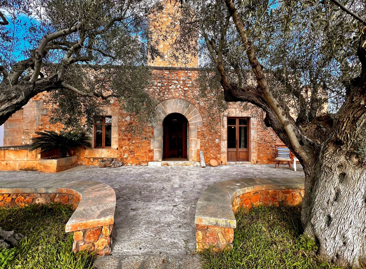 Historic stone monastery exterior with olive trees and pathways under bright blue Mediterranean skies.