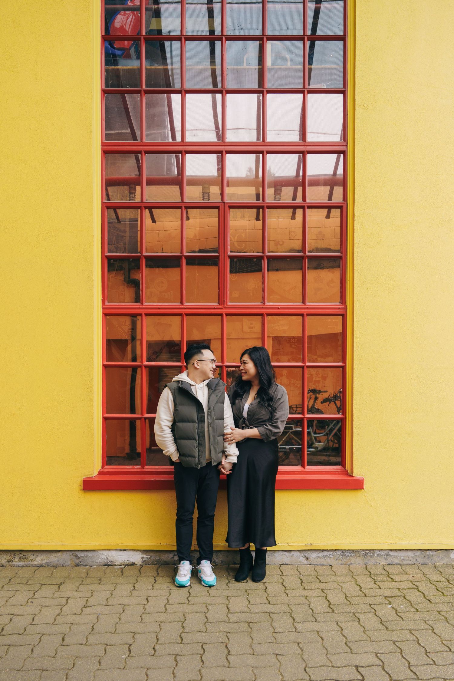 A couple shares a tender moment against a vibrant yellow wall with red-framed windows.