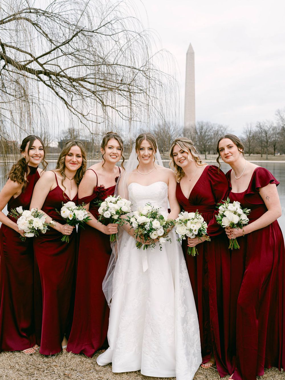 A bridal party in burgundy bridesmaid dresses and white wedding gown pose together against a winter landscape.