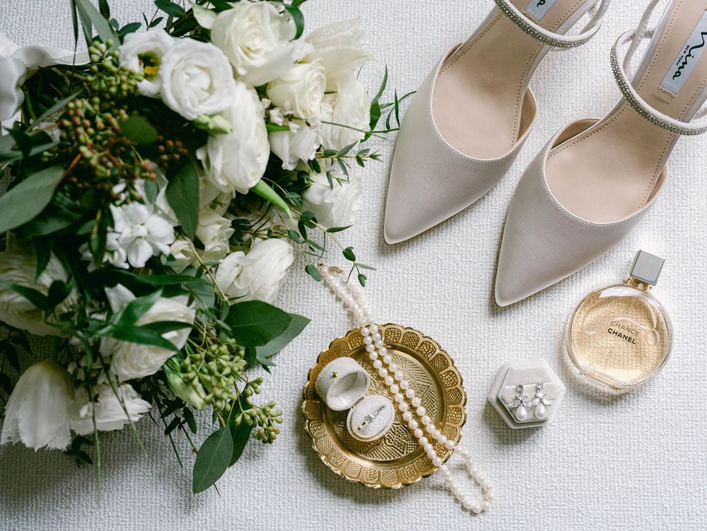 Wedding accessories including white bridal shoes, bouquet, and gold decorative elements arranged on light background.