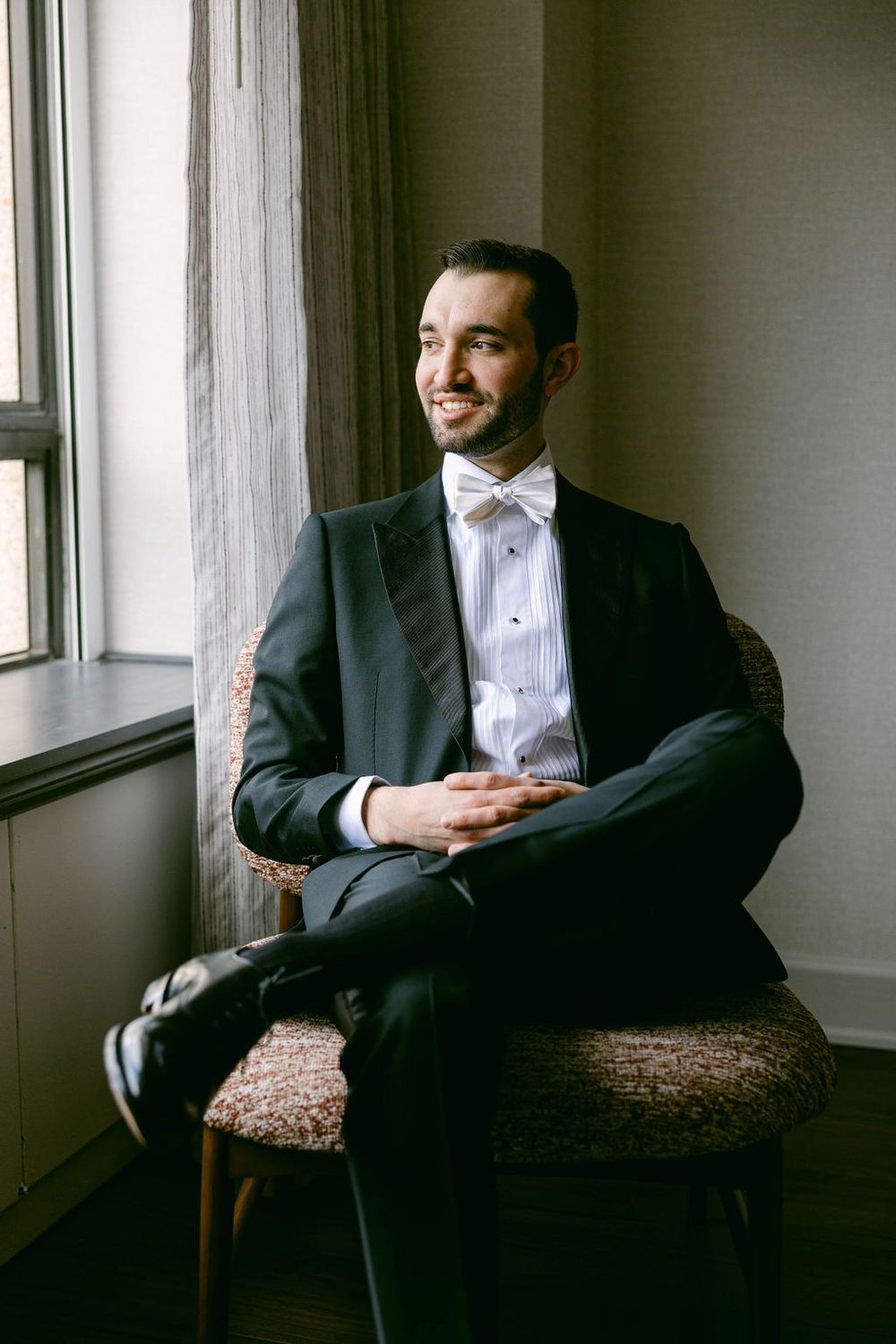 A person in a tuxedo sits contemplatively in a chair by a window in a minimalist room.