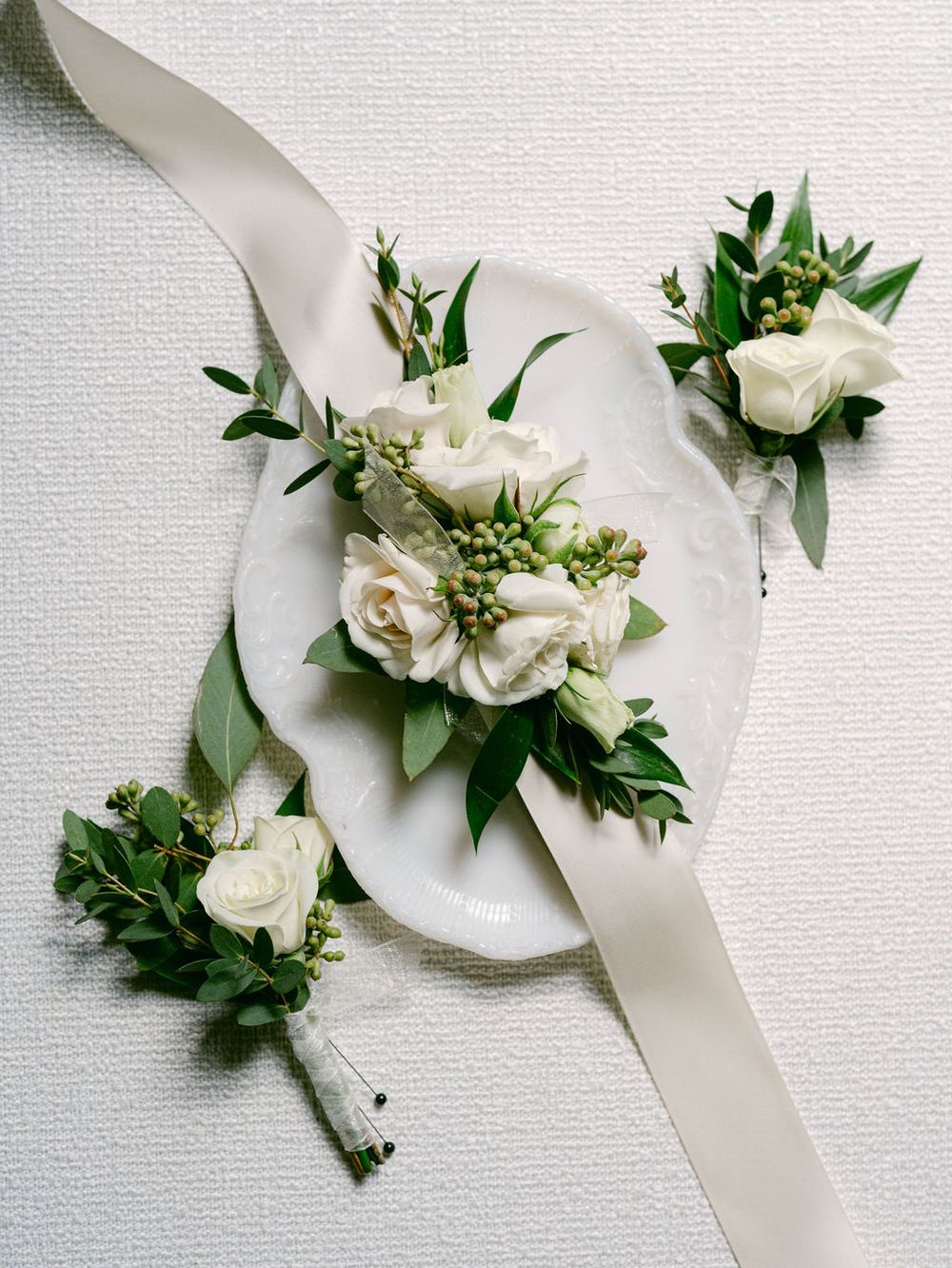 White roses and greenery arranged on silk ribbons create elegant wedding corsages on a light background.