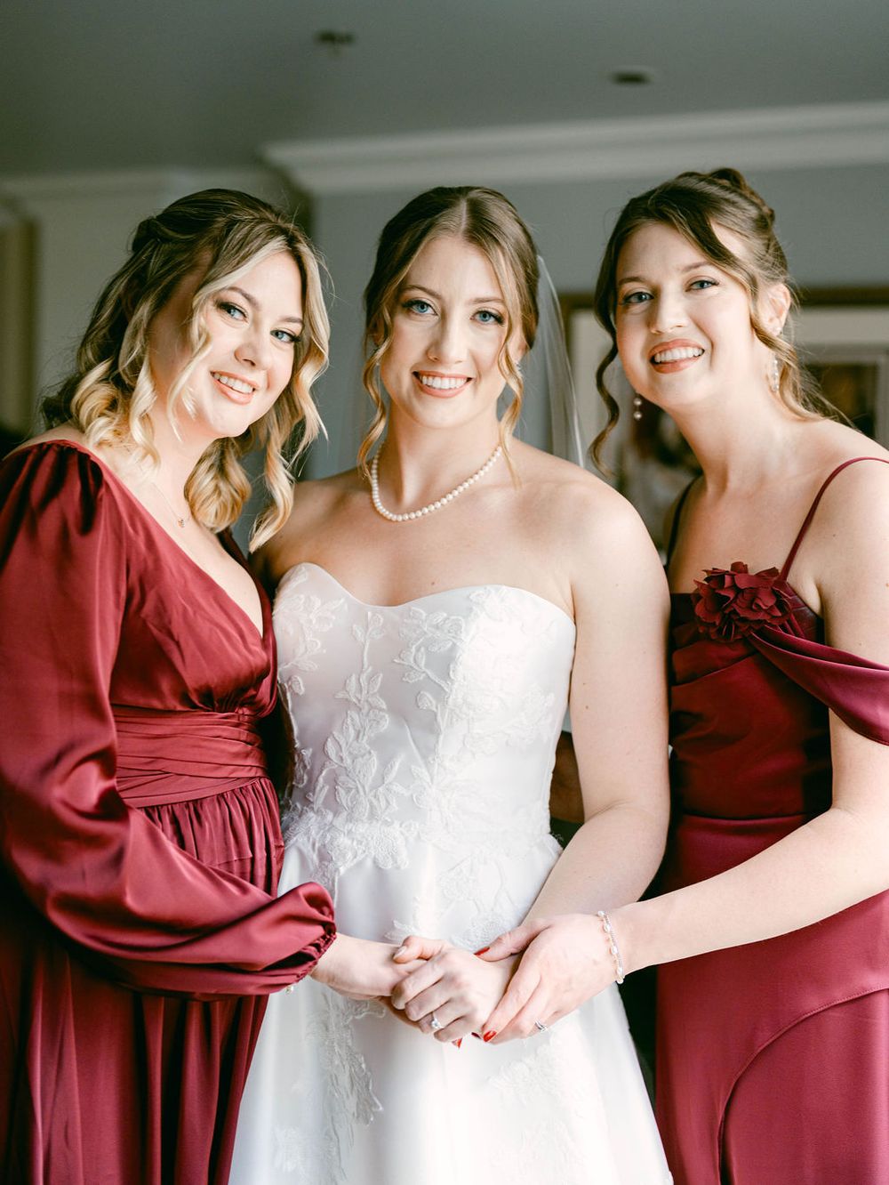 Three women in burgundy dresses and a white wedding gown pose together for a bridal party portrait.
