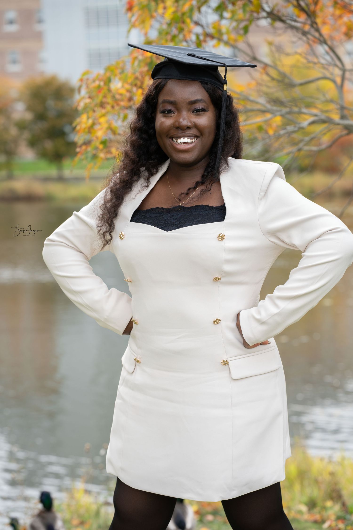 Graduate in white blazer dress and cap poses confidently by autumn lake with fall foliage in background.