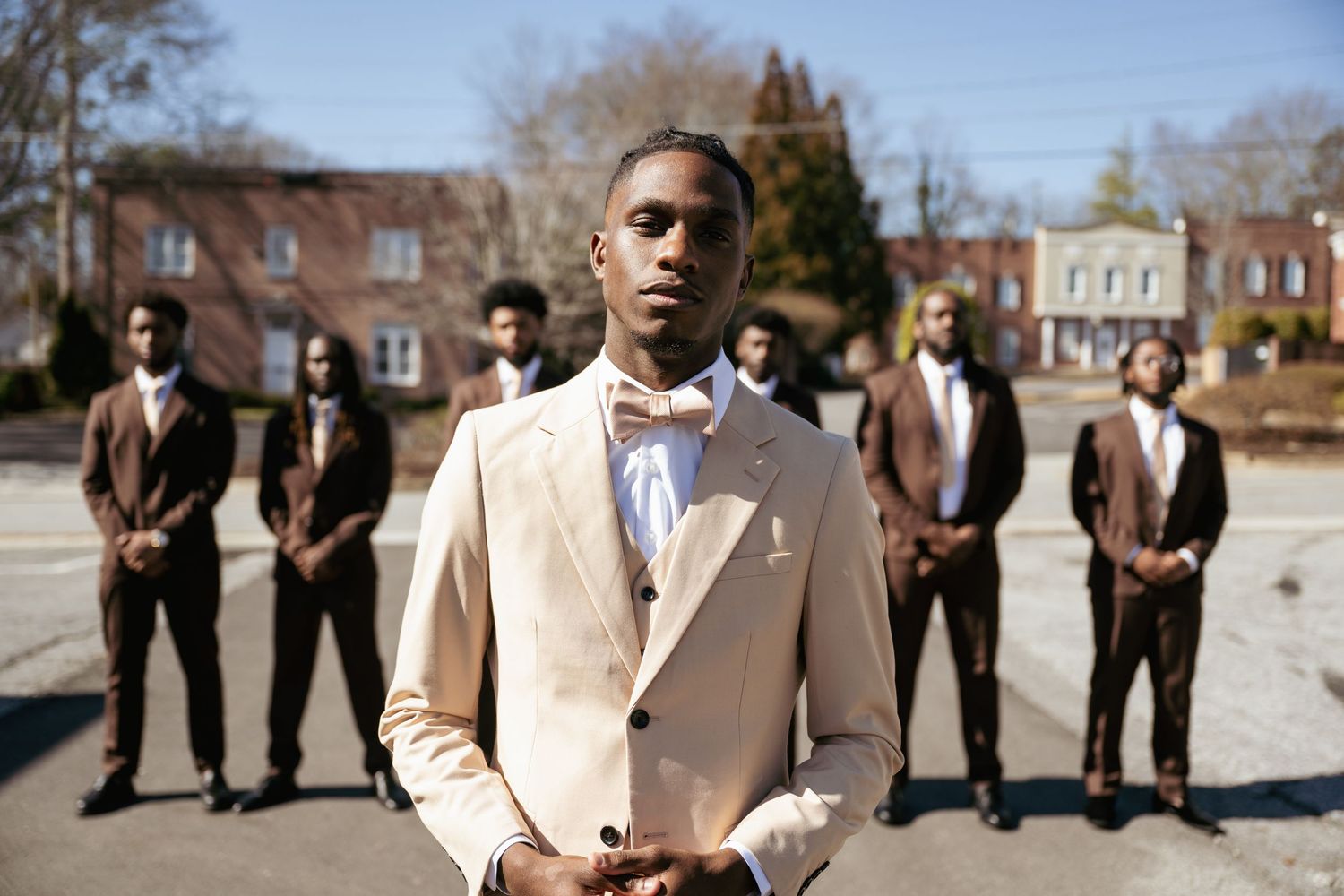 A wedding party poses outdoors with the groom in a cream-colored suit standing in front.
