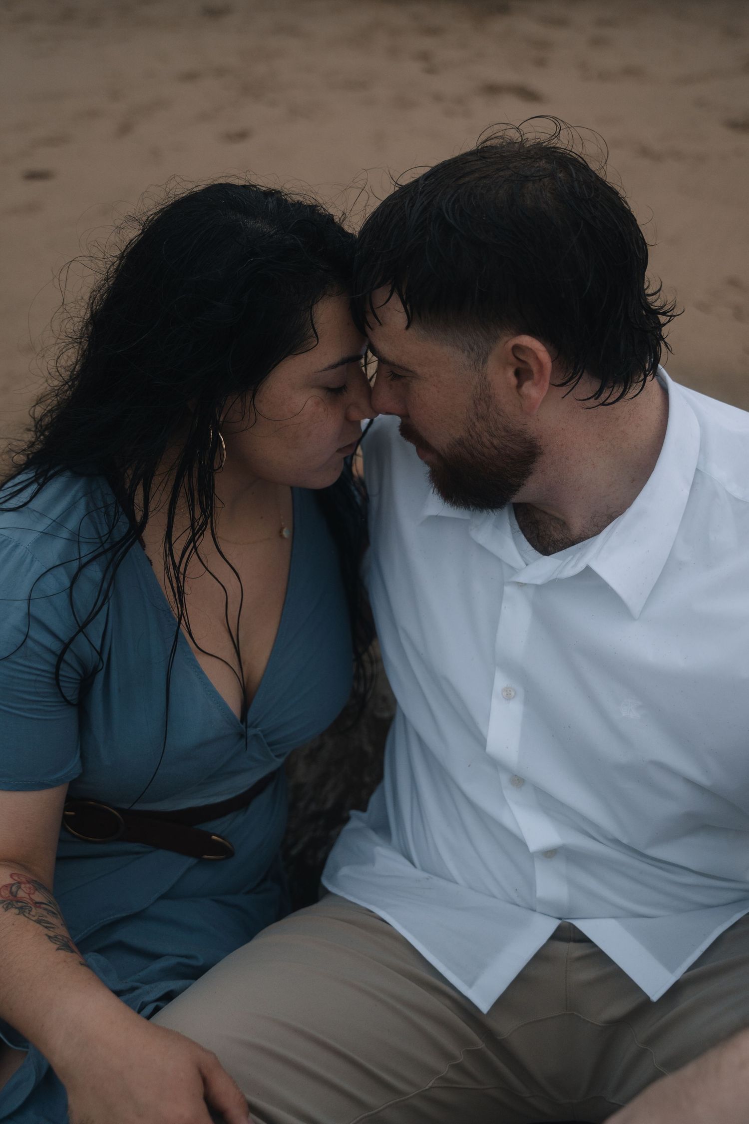 Intimate couple sharing tender moment on sandy beach, wearing blue dress and white shirt.