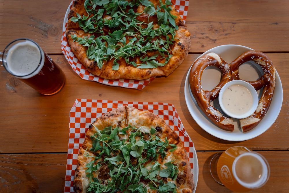 Table spread with pizza, pretzel, and craft beers on wooden surface.