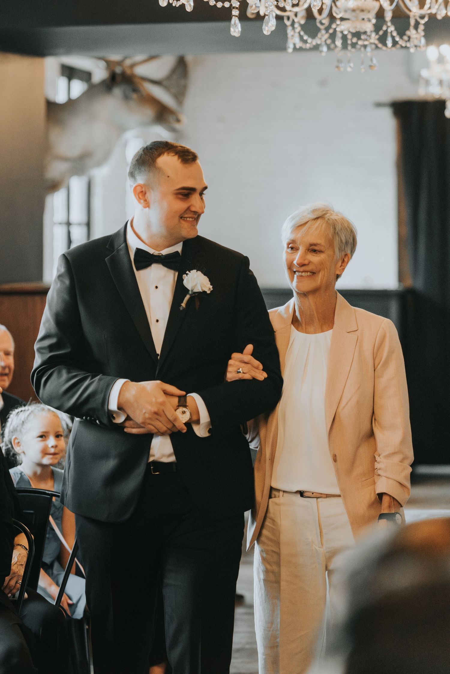 Wedding ceremony processional in an elegant venue with a chandelier overhead and two people walking arm in arm.