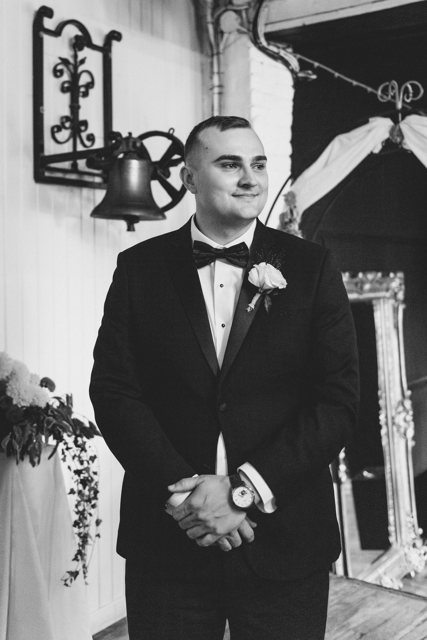Black and white photograph of a groom in a classic tuxedo with bow tie and boutonniere at a formal wedding ceremony.