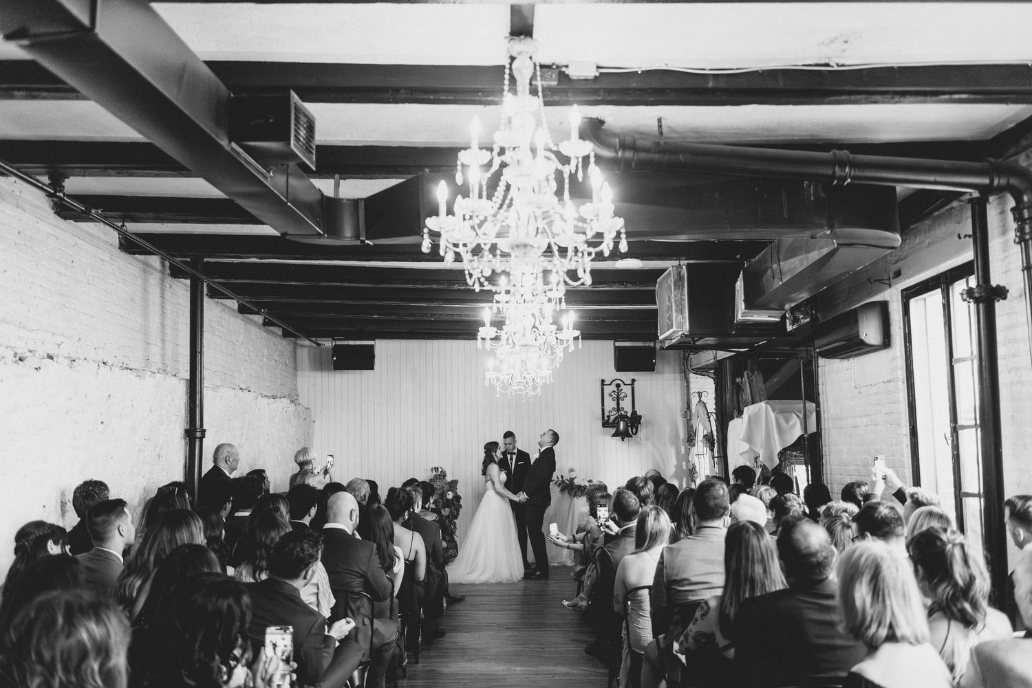 A black and white wedding ceremony takes place in an elegant industrial venue with exposed beams and a crystal chandelier overhead.