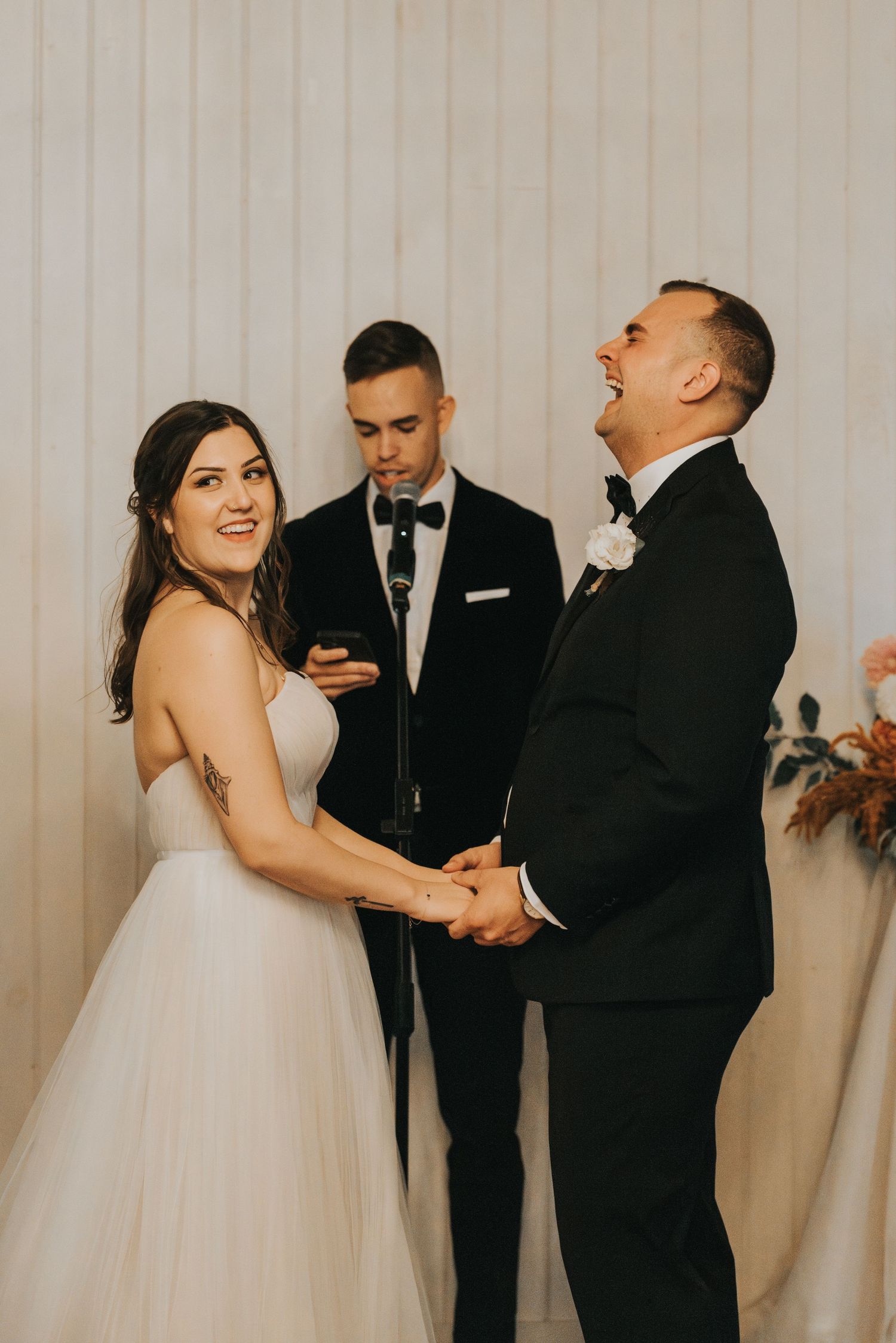 Candid moment during wedding ceremony with couple sharing laughter and holding hands while officiant reads.
