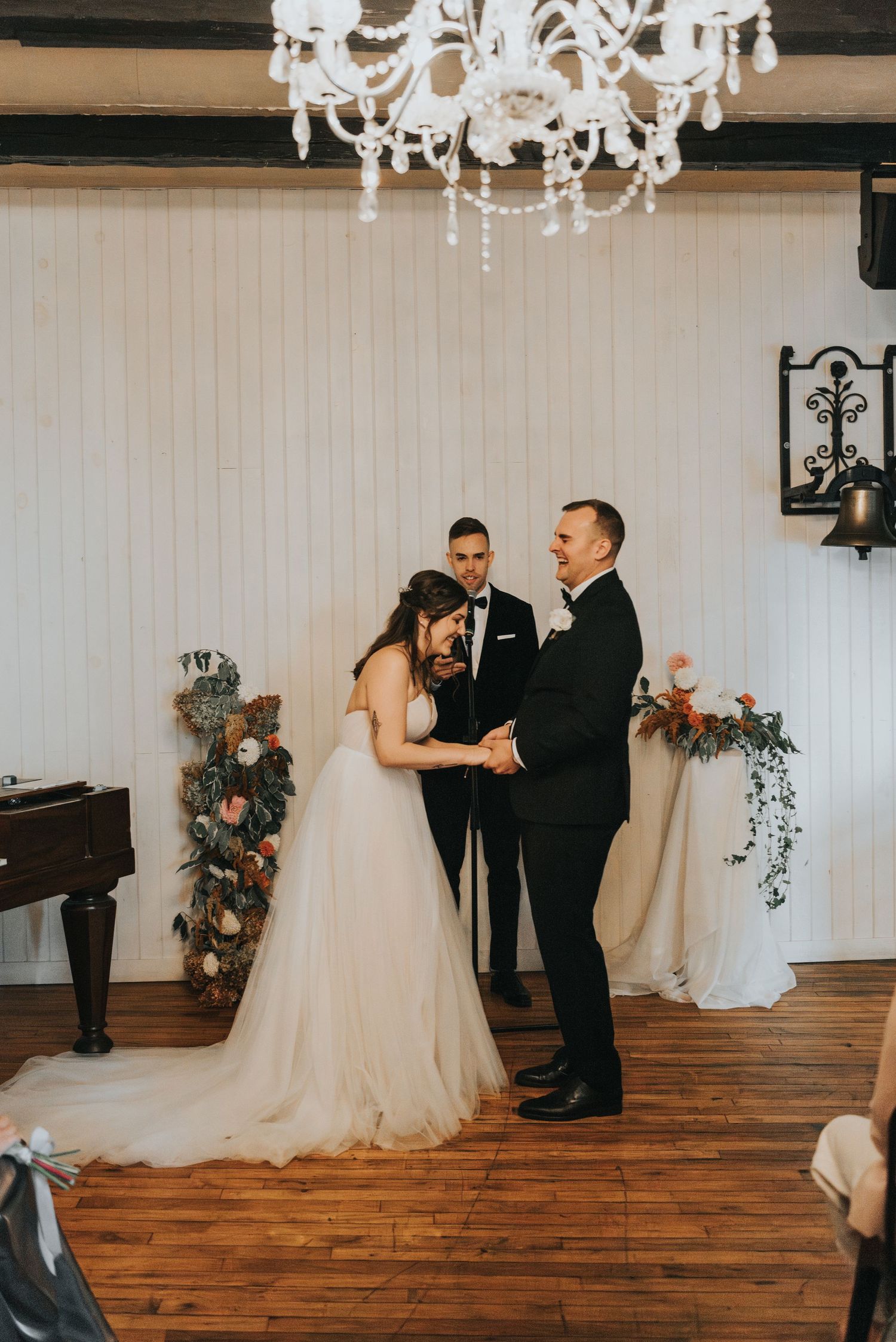 Wedding ceremony inside rustic venue with crystal chandelier, wooden floors and elegant floral decorations.