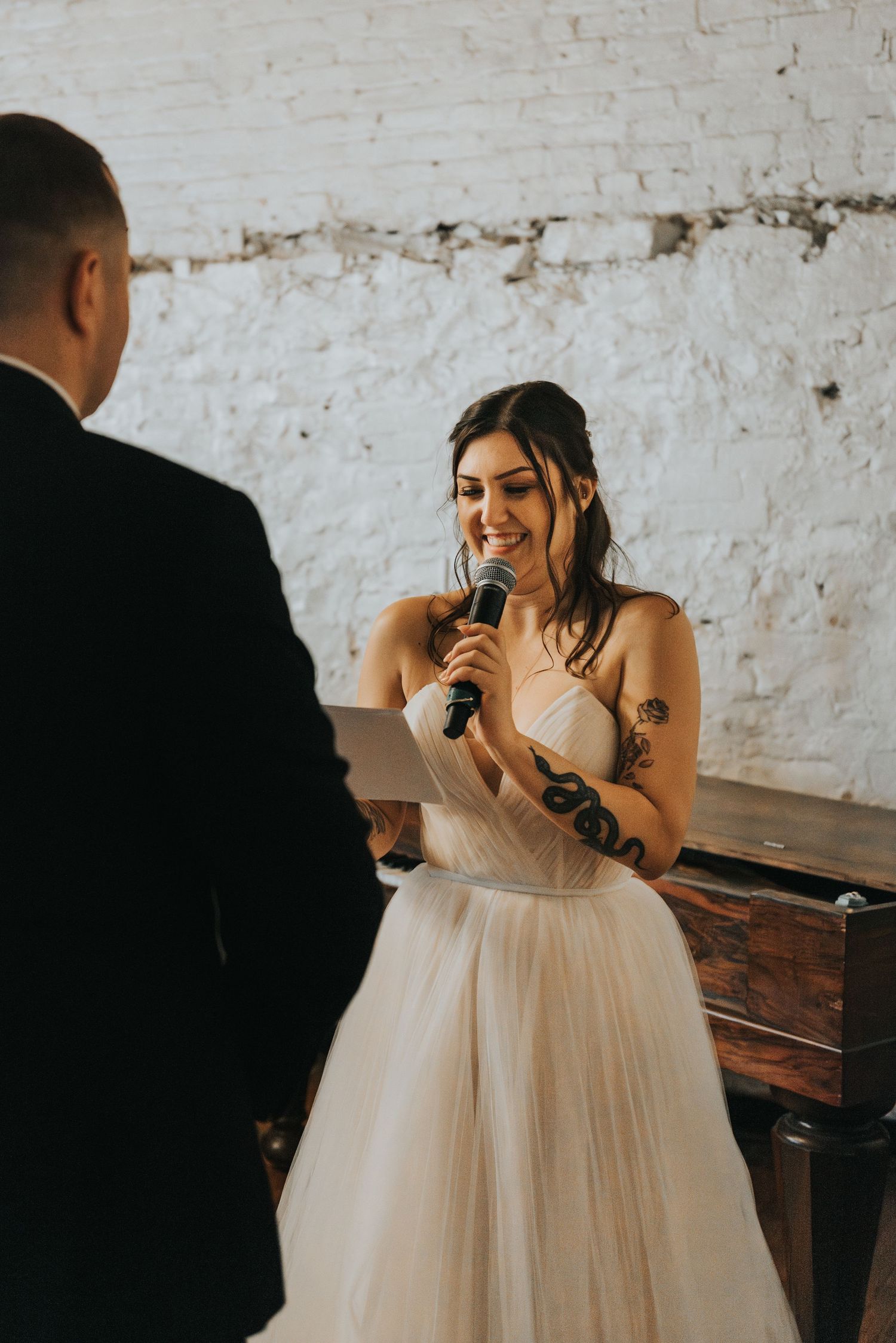 A bride in a flowy white gown reads from papers while giving a speech at her wedding ceremony.