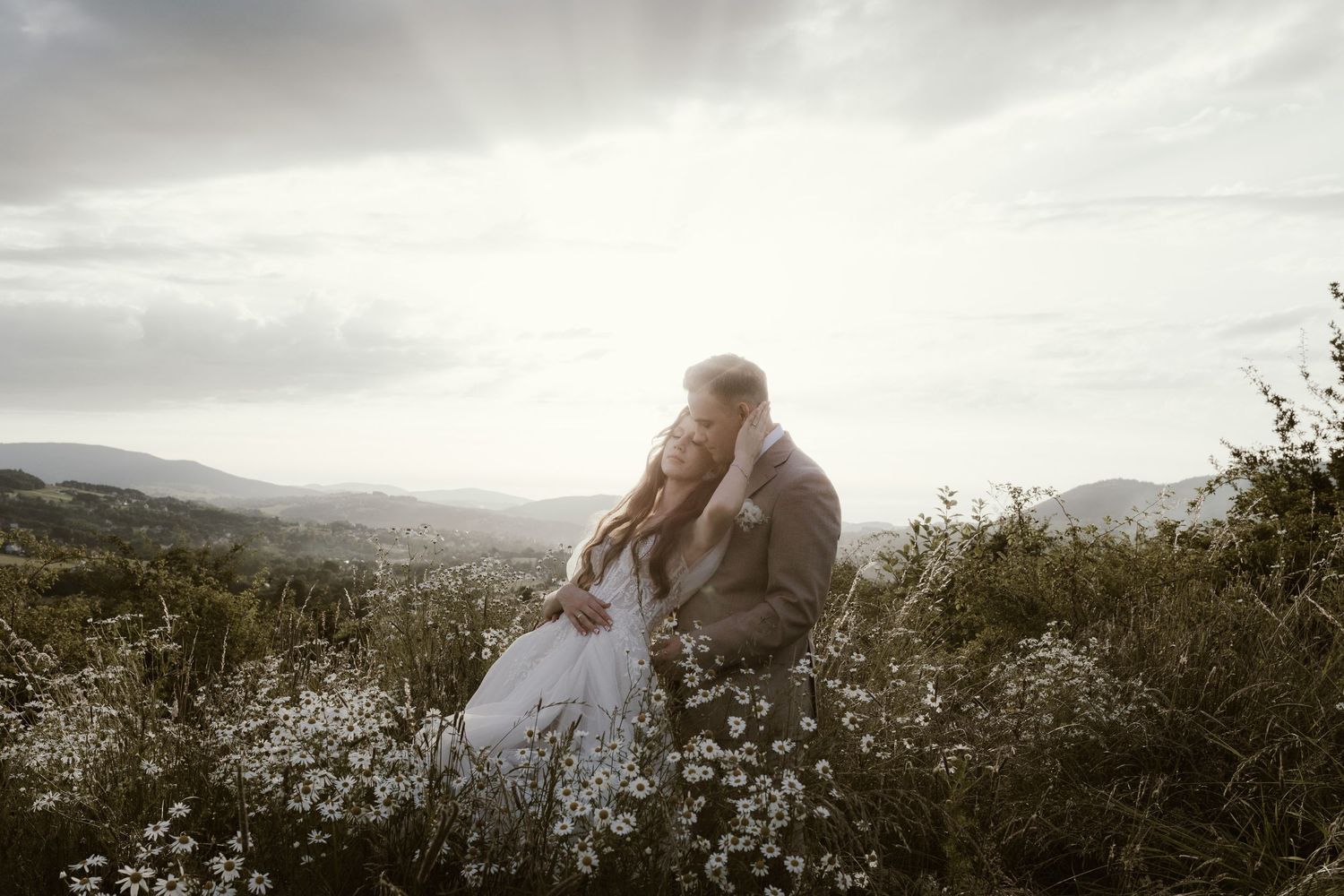 Two people in wedding attire embrace while sitting in a mountainside meadow at sunset against a cloudy sky.