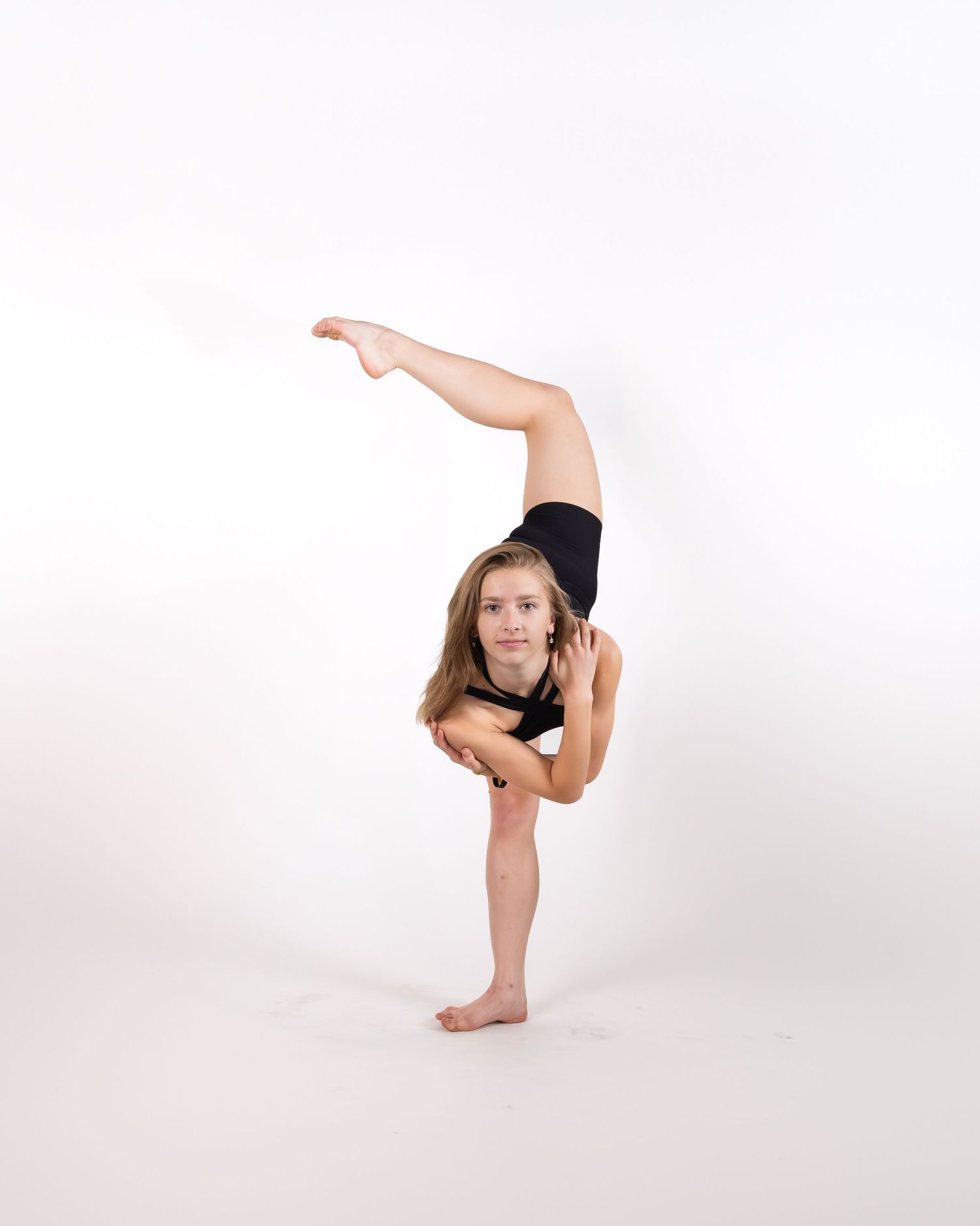 Gymnast in black leotard performing a standing split pose against white background.
