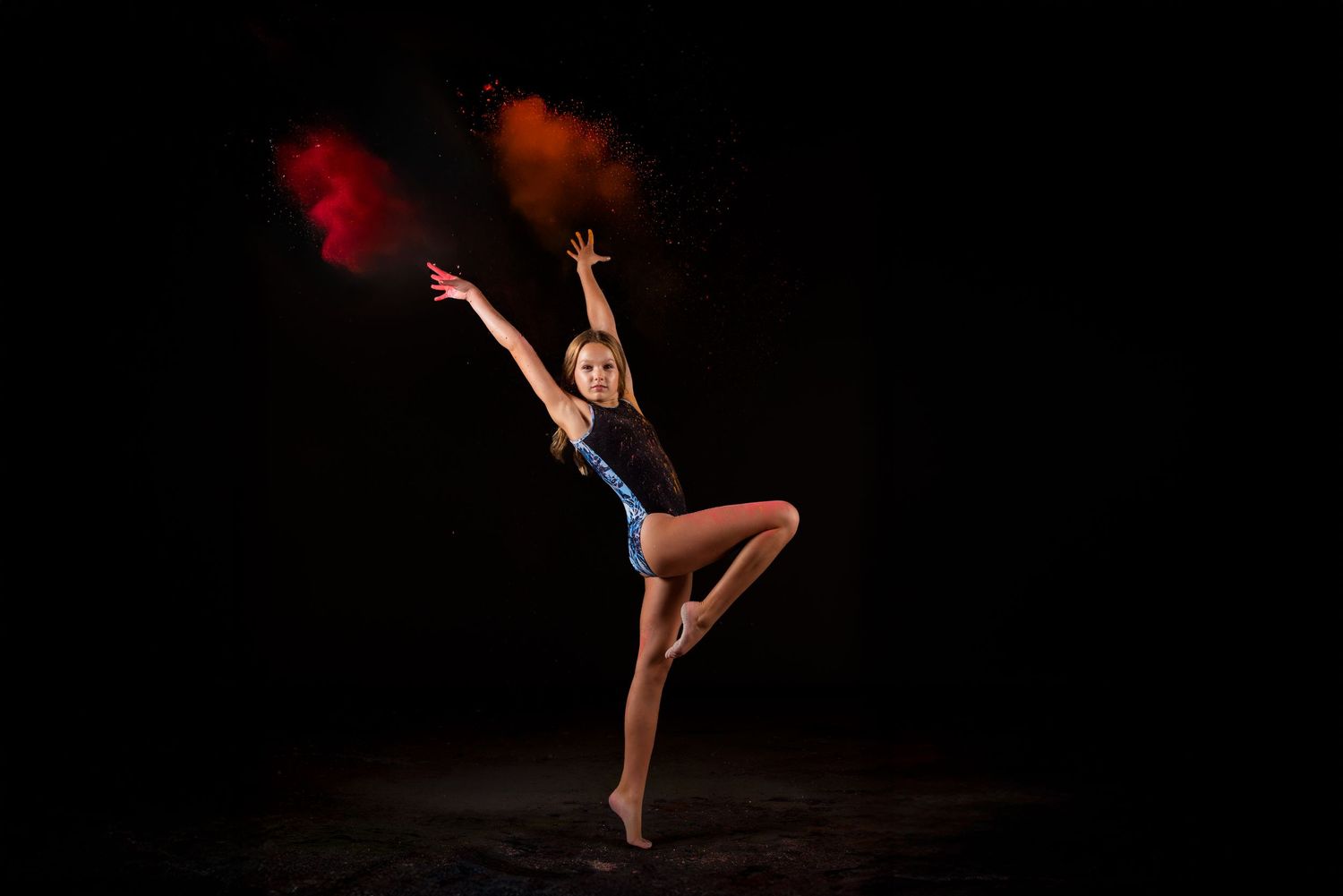 A dancer performs a graceful leap against a dark backdrop while holding colorful red powder that creates a dramatic effect.
