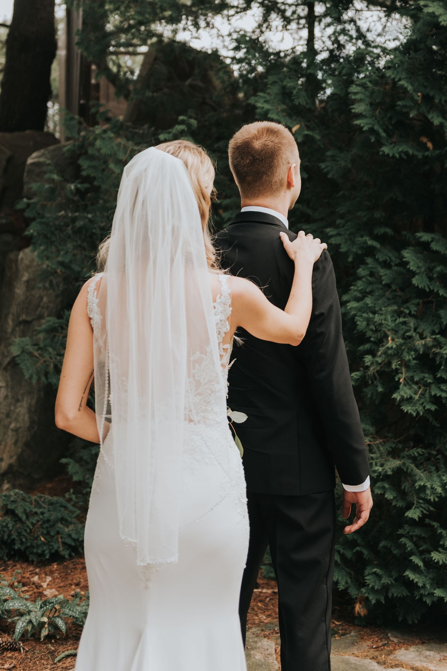 A bride in a fitted lace gown and veil places her hand on the groom's shoulder as they stand amid garden greenery.