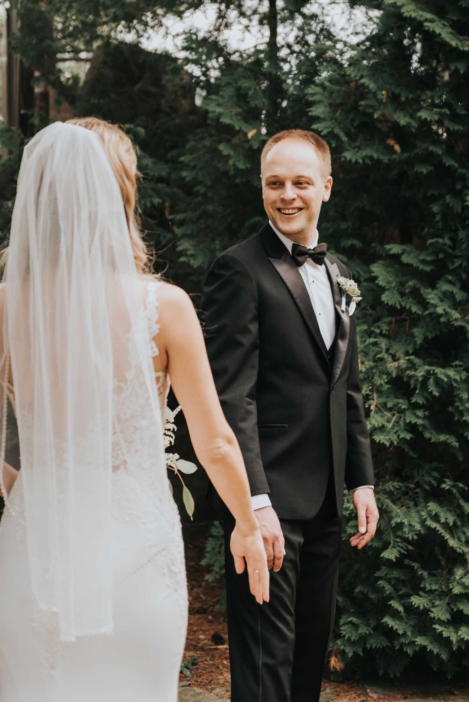A romantic wedding first look moment captured outdoors with greenery and trees in the background.