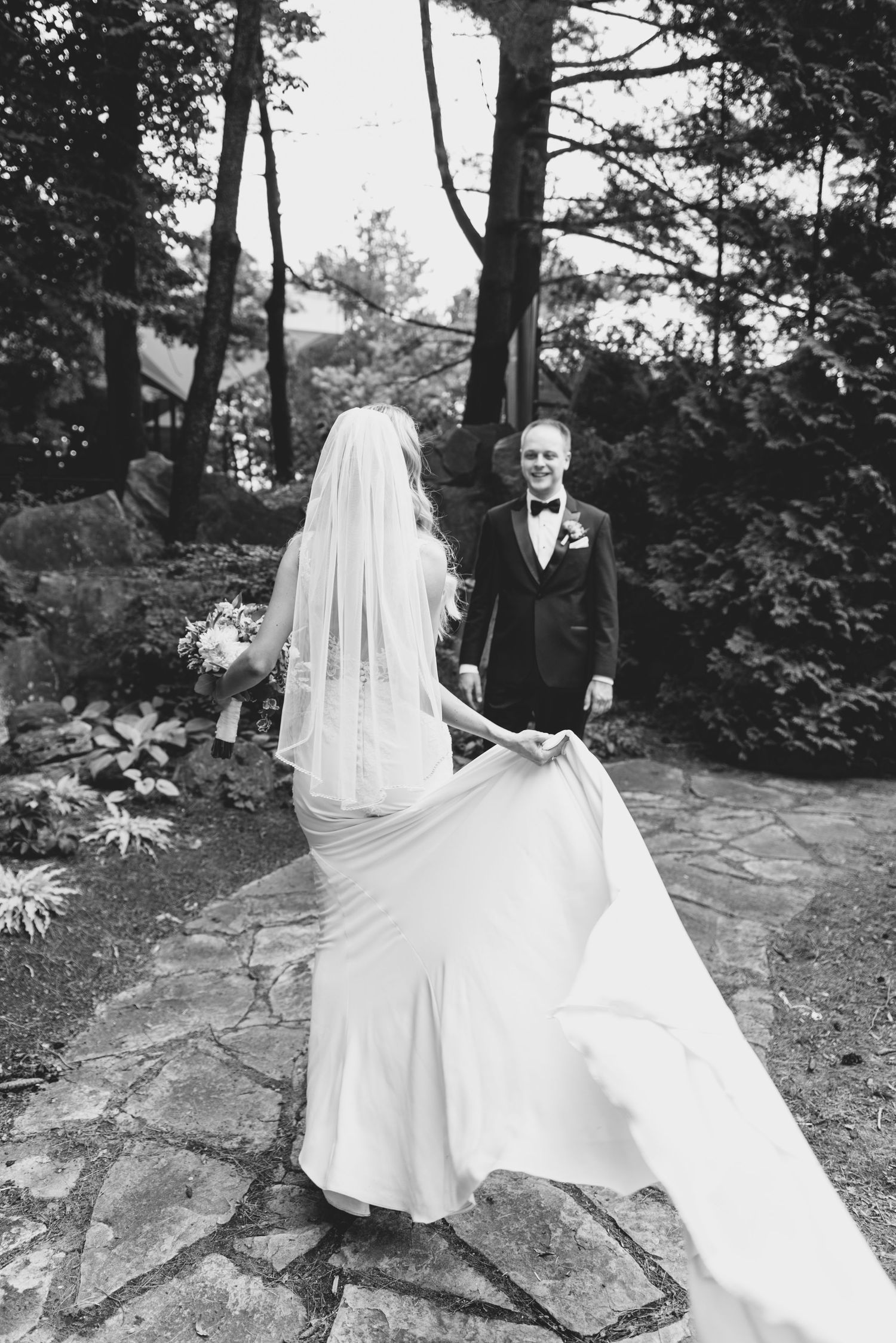 A bride in a flowing white dress spins on stone steps surrounded by forest scenery.