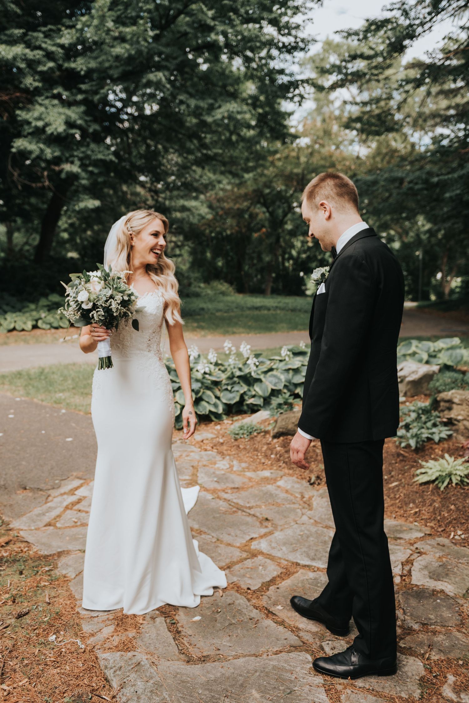 A bride in a white fitted gown and groom in black suit share a candid moment on stone path surrounded by greenery.