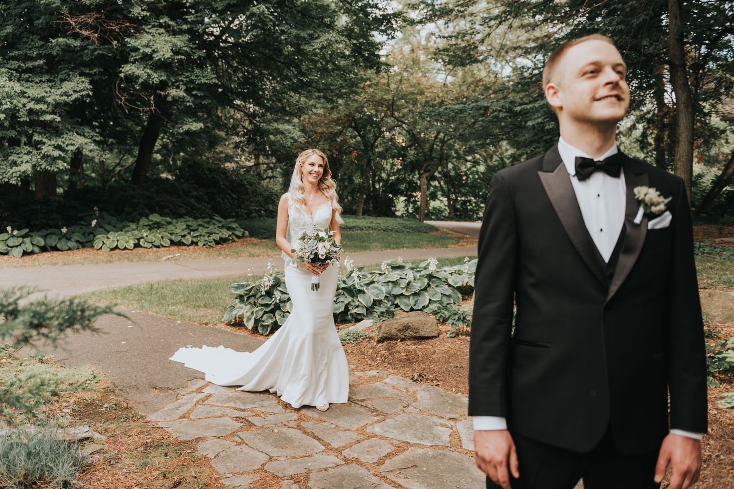 A couple in wedding attire during their first look moment in a garden setting with stone pathways and lush greenery.