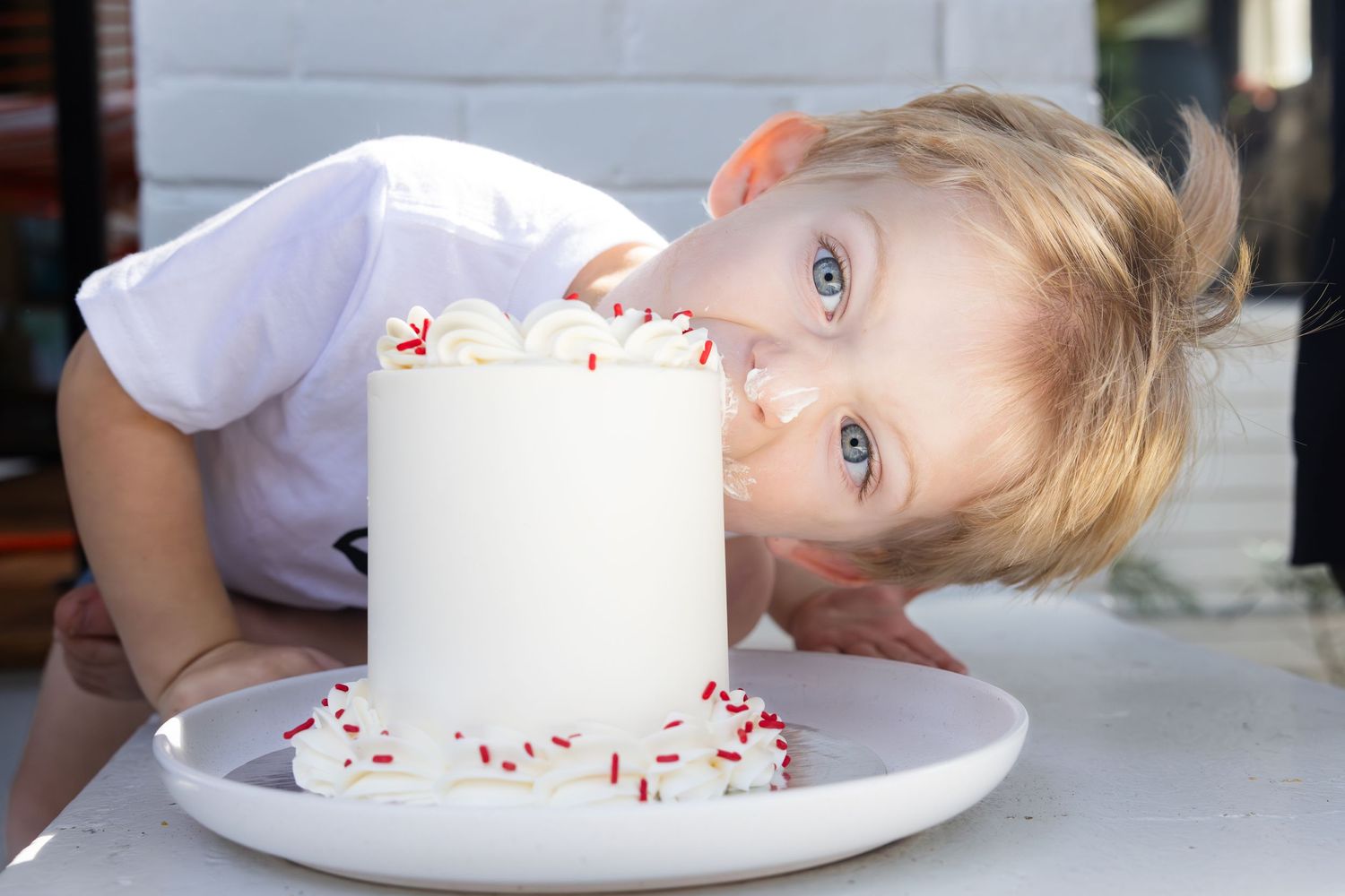 Child taking a bite from a large white candle decorated with red sprinkles on a white plate.