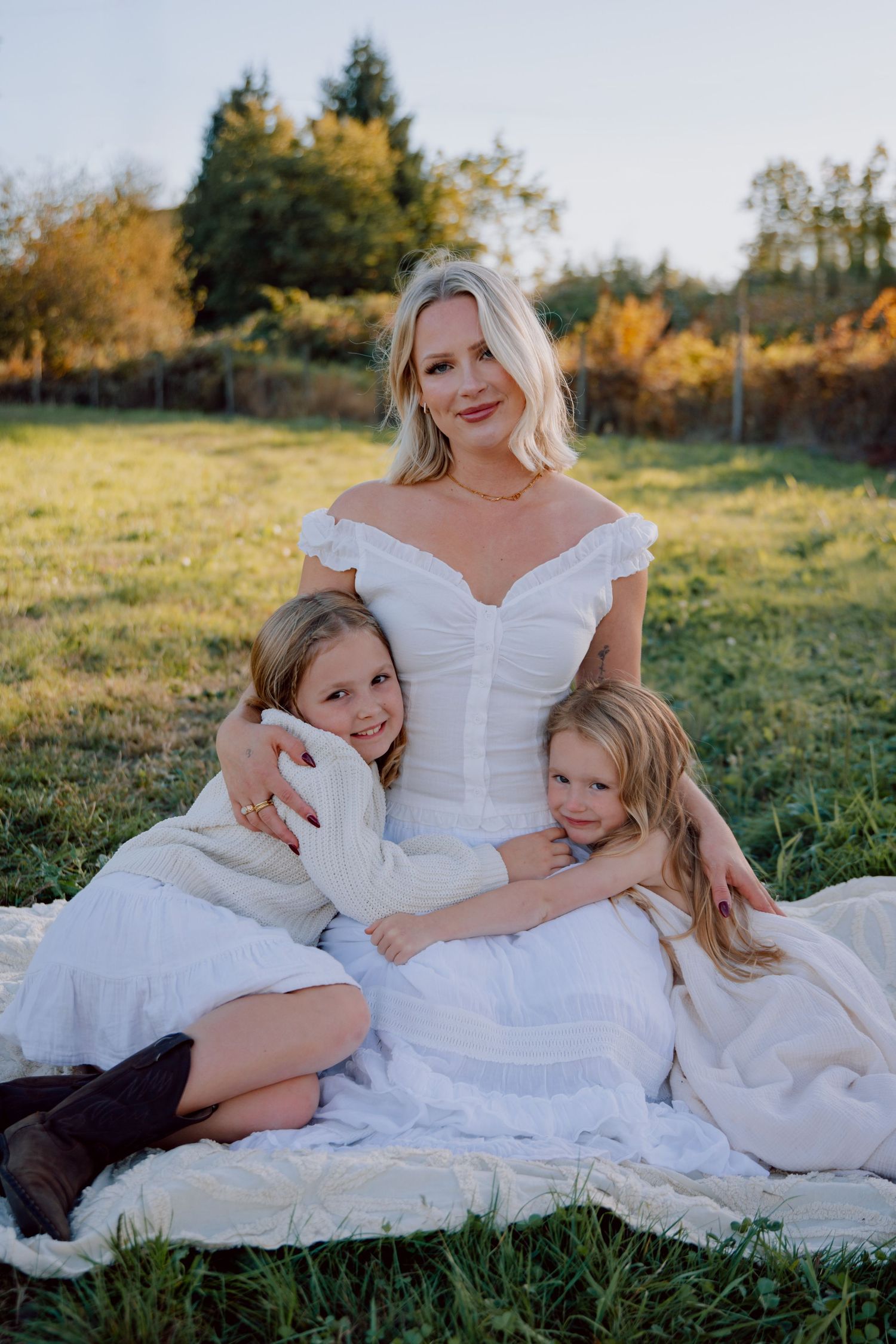 Family wearing white dresses rests together on grassy field during golden hour sunset photo session.