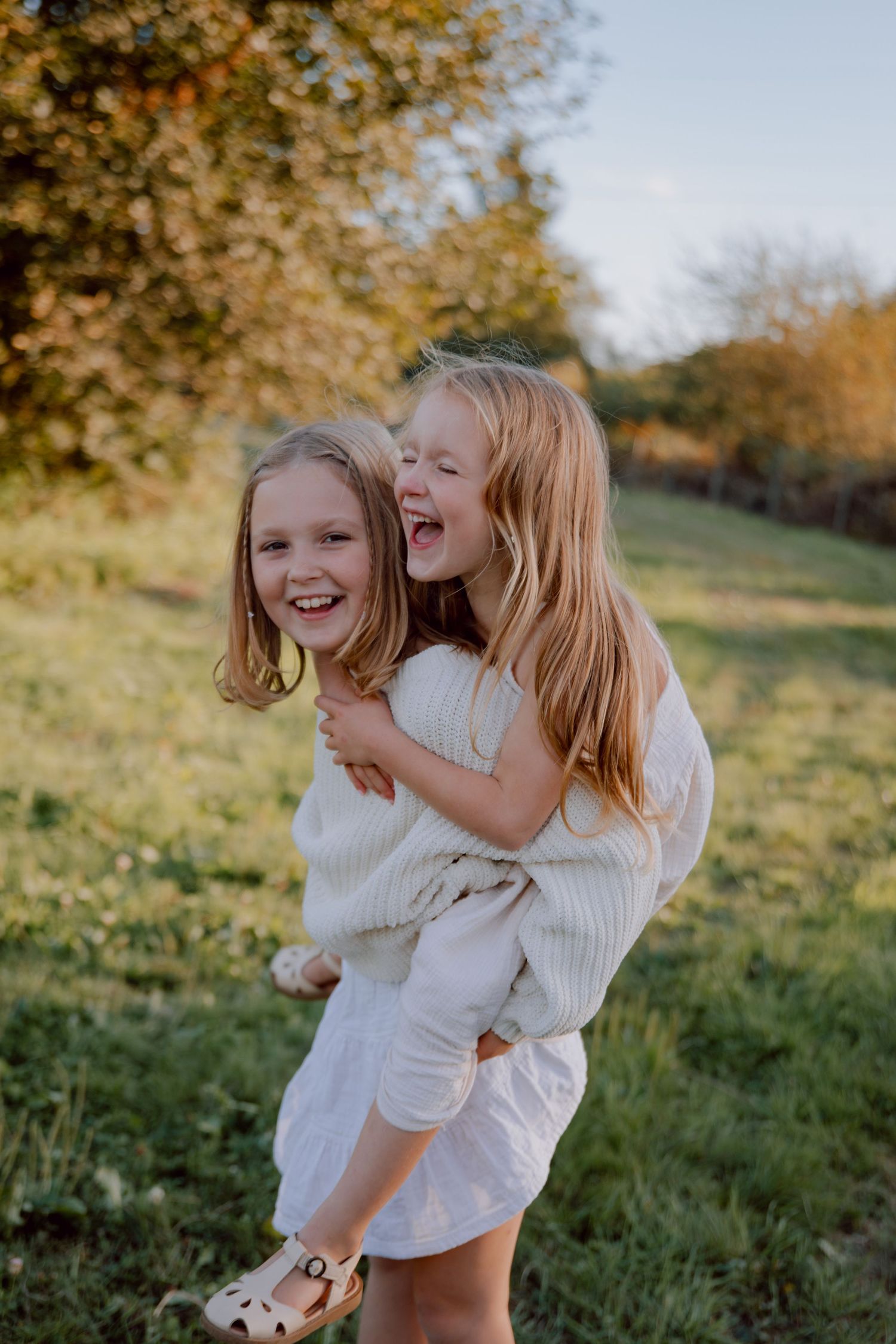 Two children in white outfits laughing and playing piggyback in a sunny autumn field with golden foliage.