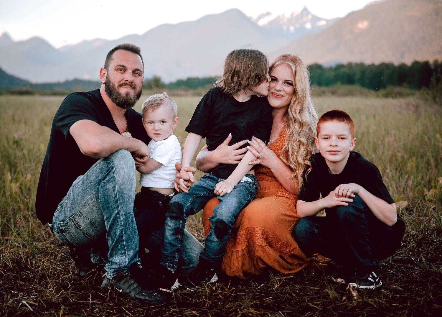 Family of five sits together in field with dramatic mountain backdrop at sunset.