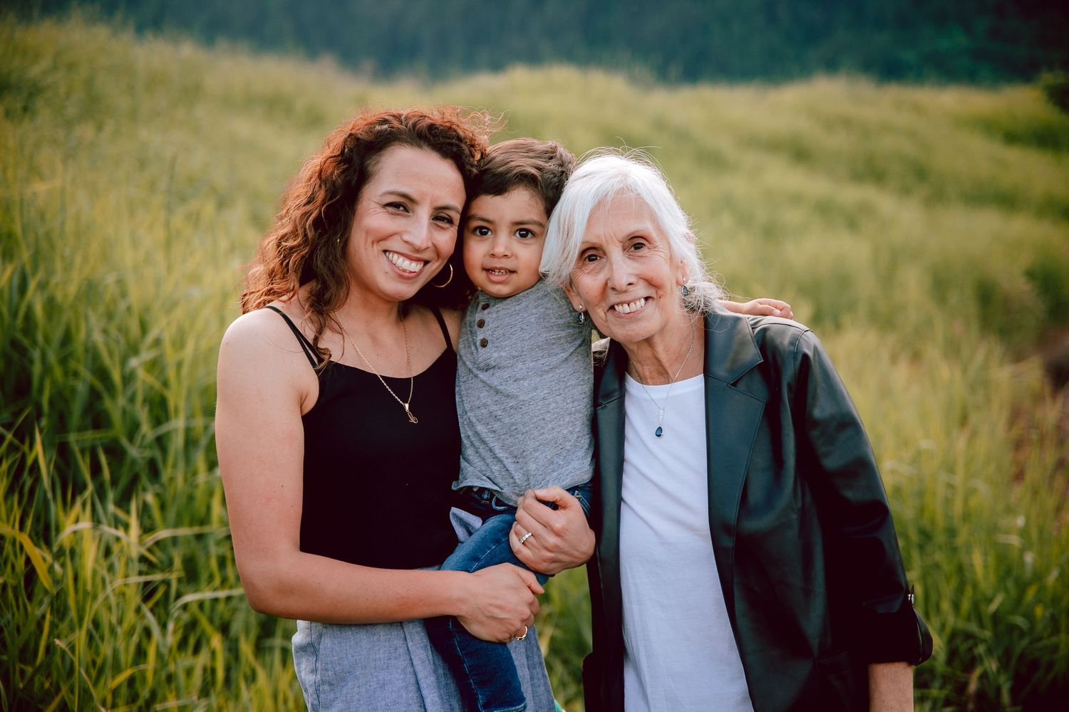 Three generations pose together in a grassy field with mountains in the background.