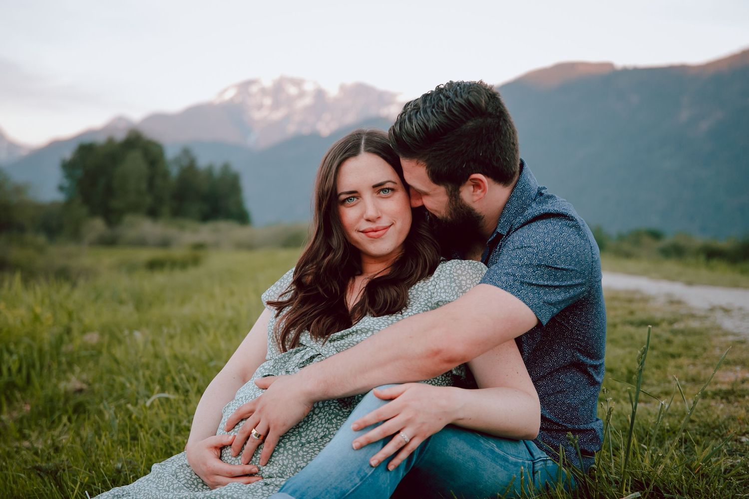 A couple shares an intimate embrace while sitting in a meadow with mountain peaks in the background at sunset.