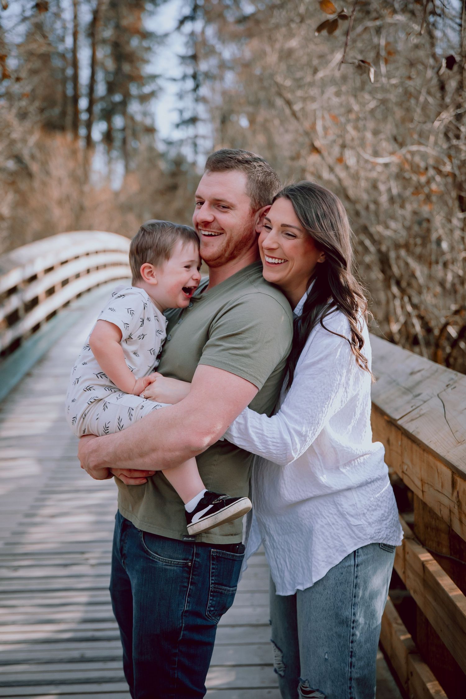 A happy family of three sharing a joyful moment together on a wooden bridge surrounded by trees.