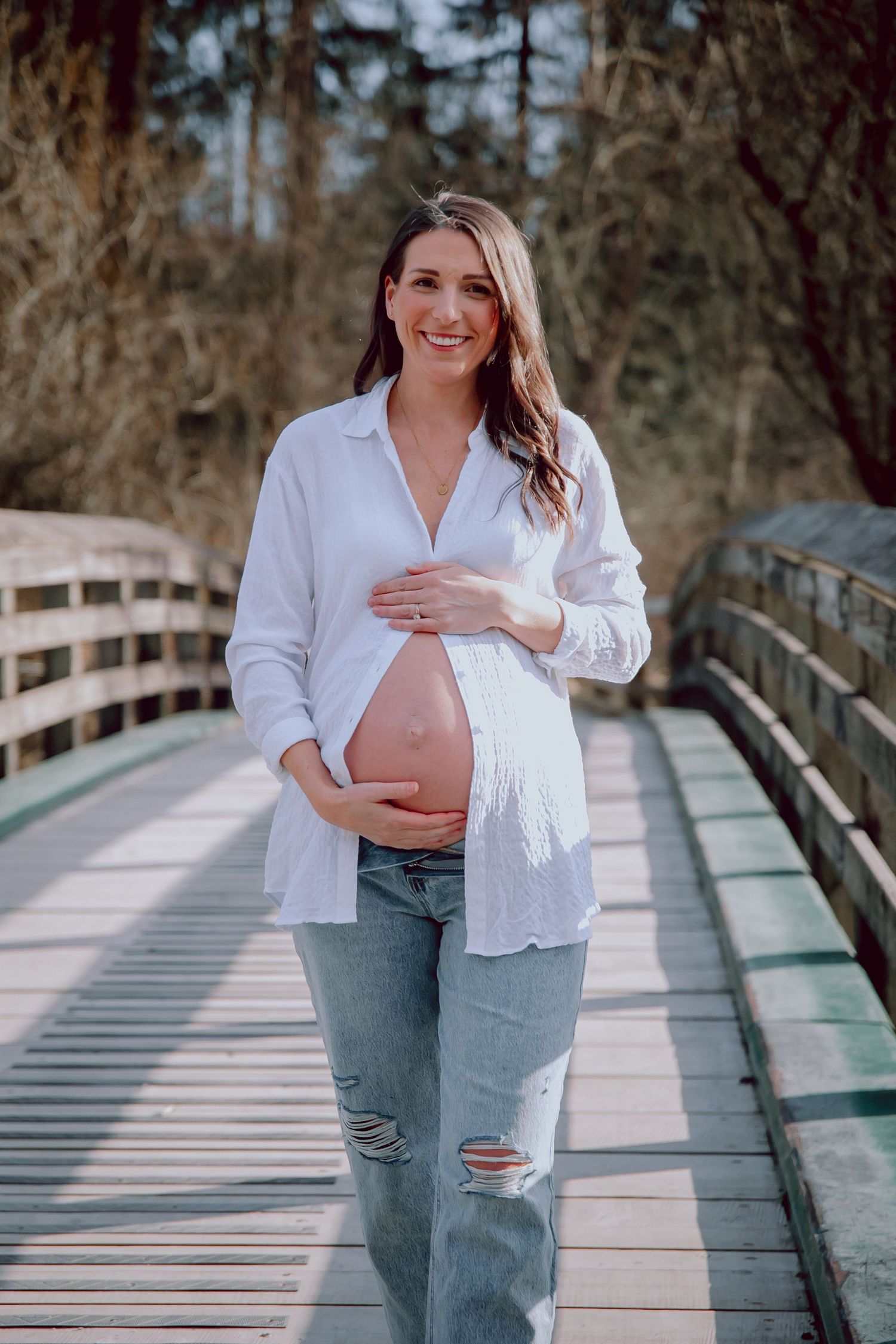 Expectant mother in white shirt walking on wooden bridge pathway while holding her baby bump.