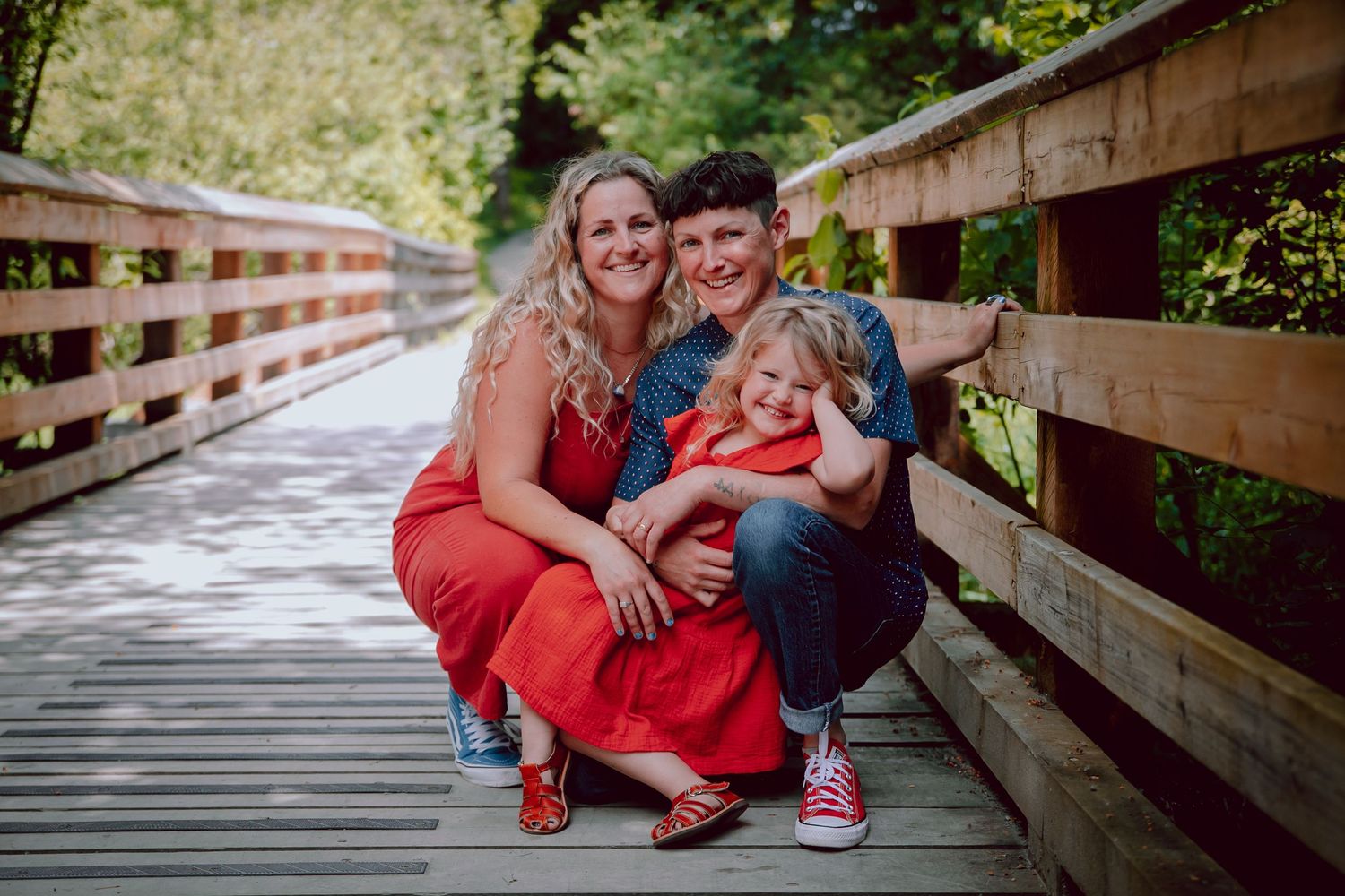 Family of three poses together on a wooden bridge walkway wearing casual clothes.