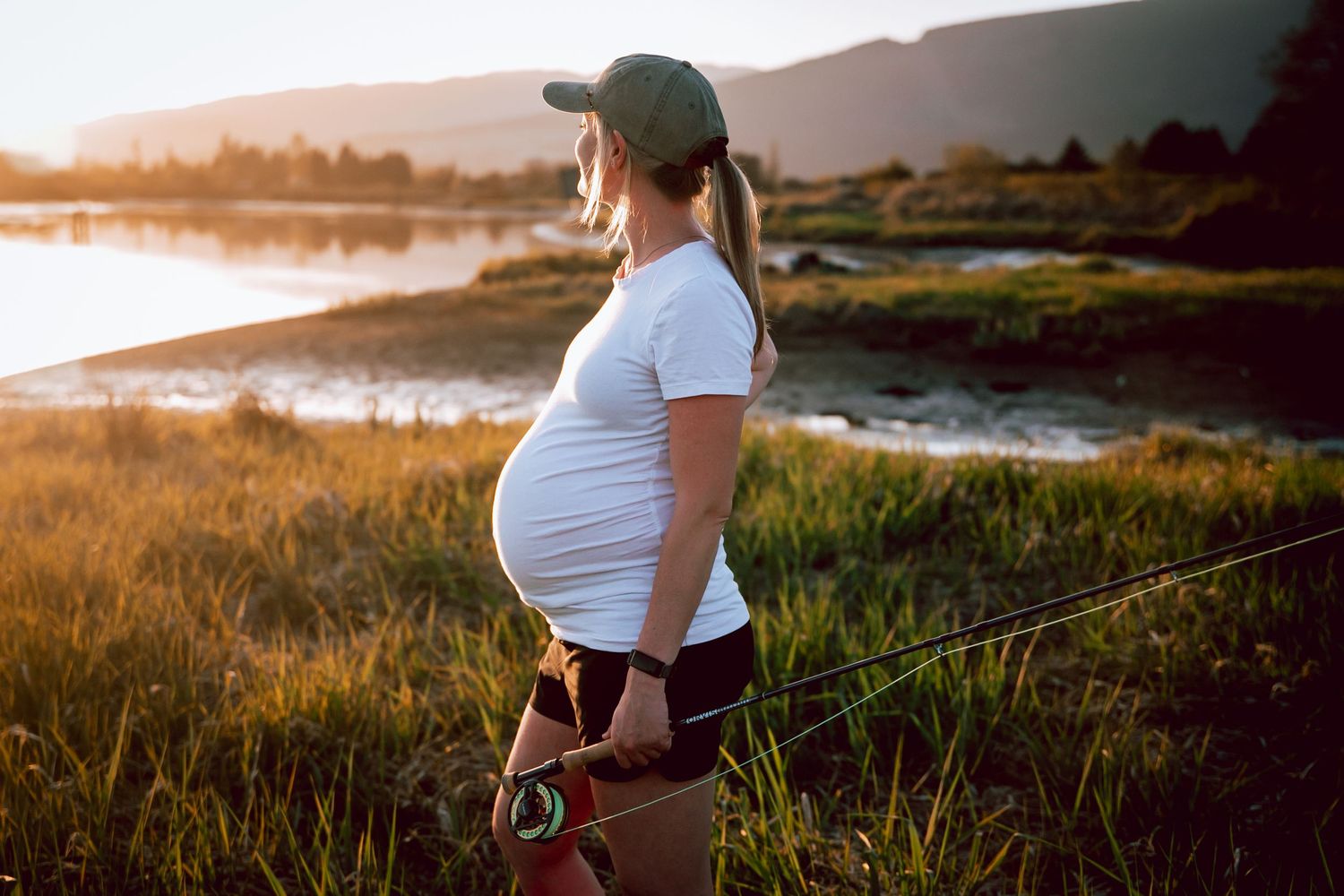 An expectant mother standing by a lake during golden hour wearing white and looking at her baby bump.