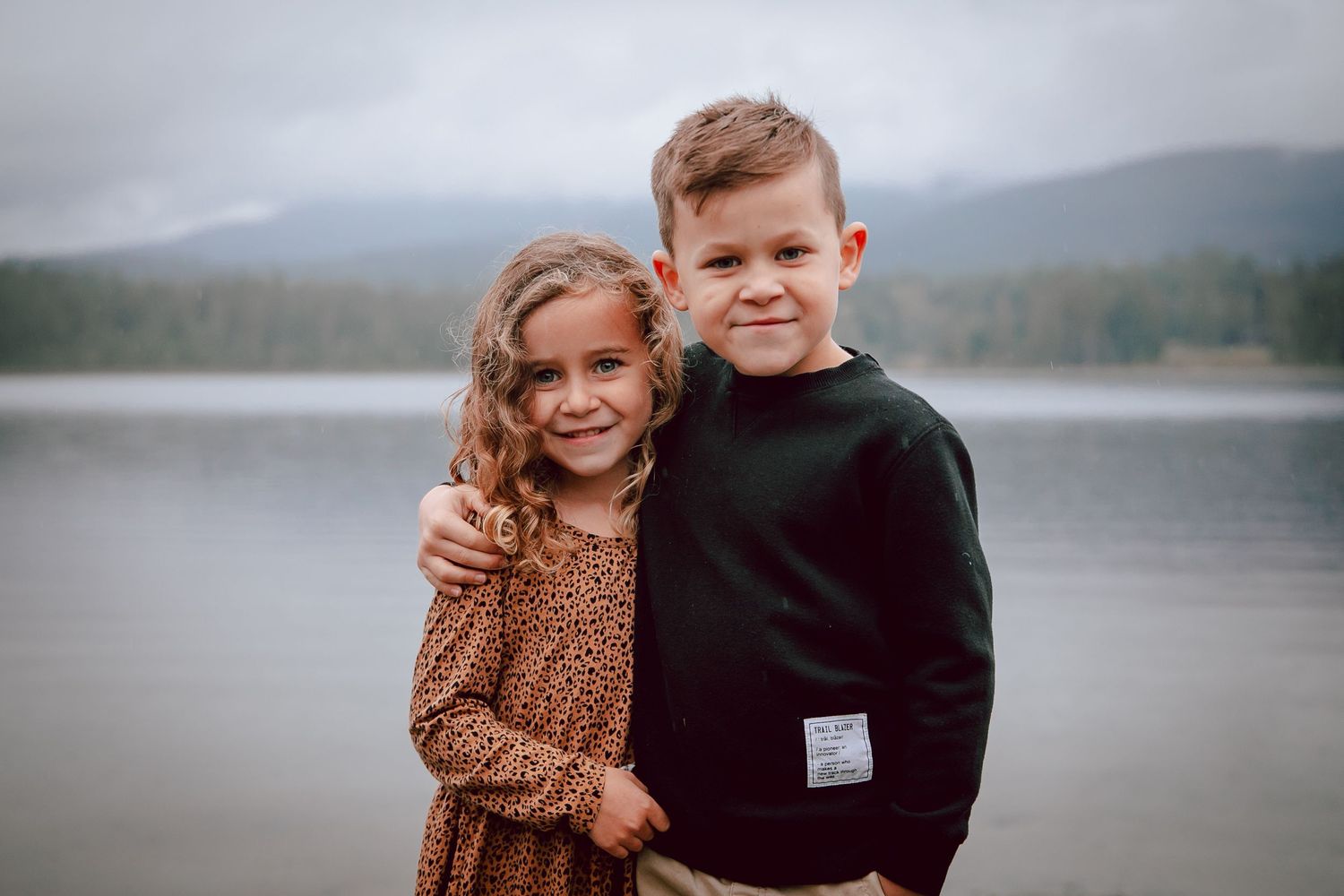 Two children stand close together smiling with a lake and mountains behind them.