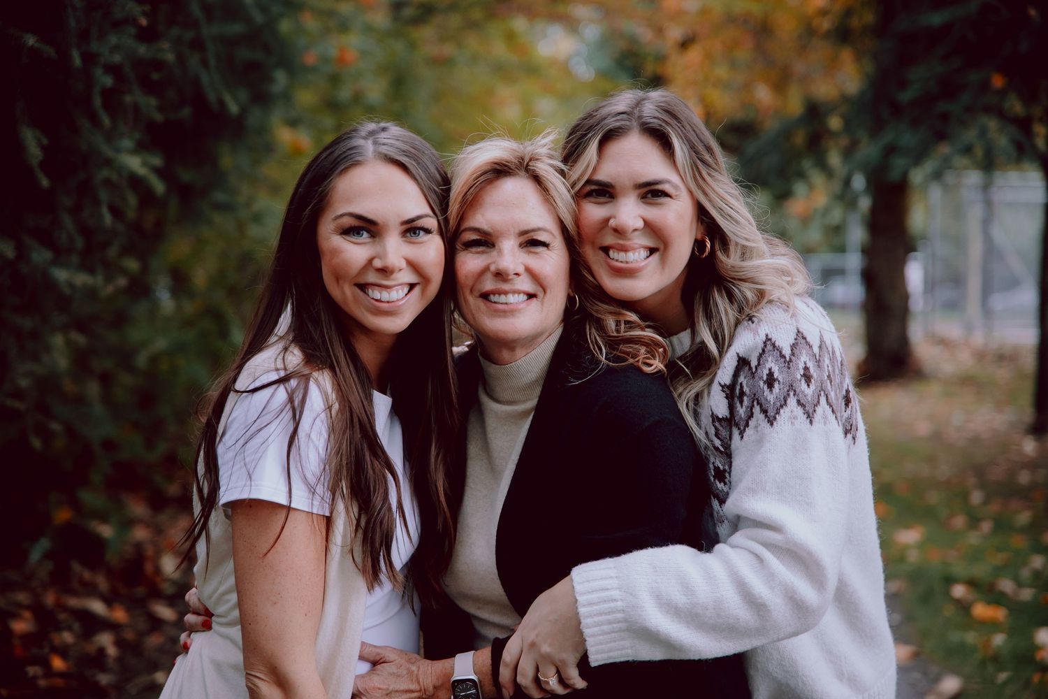 Three people share an intimate embrace while smiling outdoors during autumn.