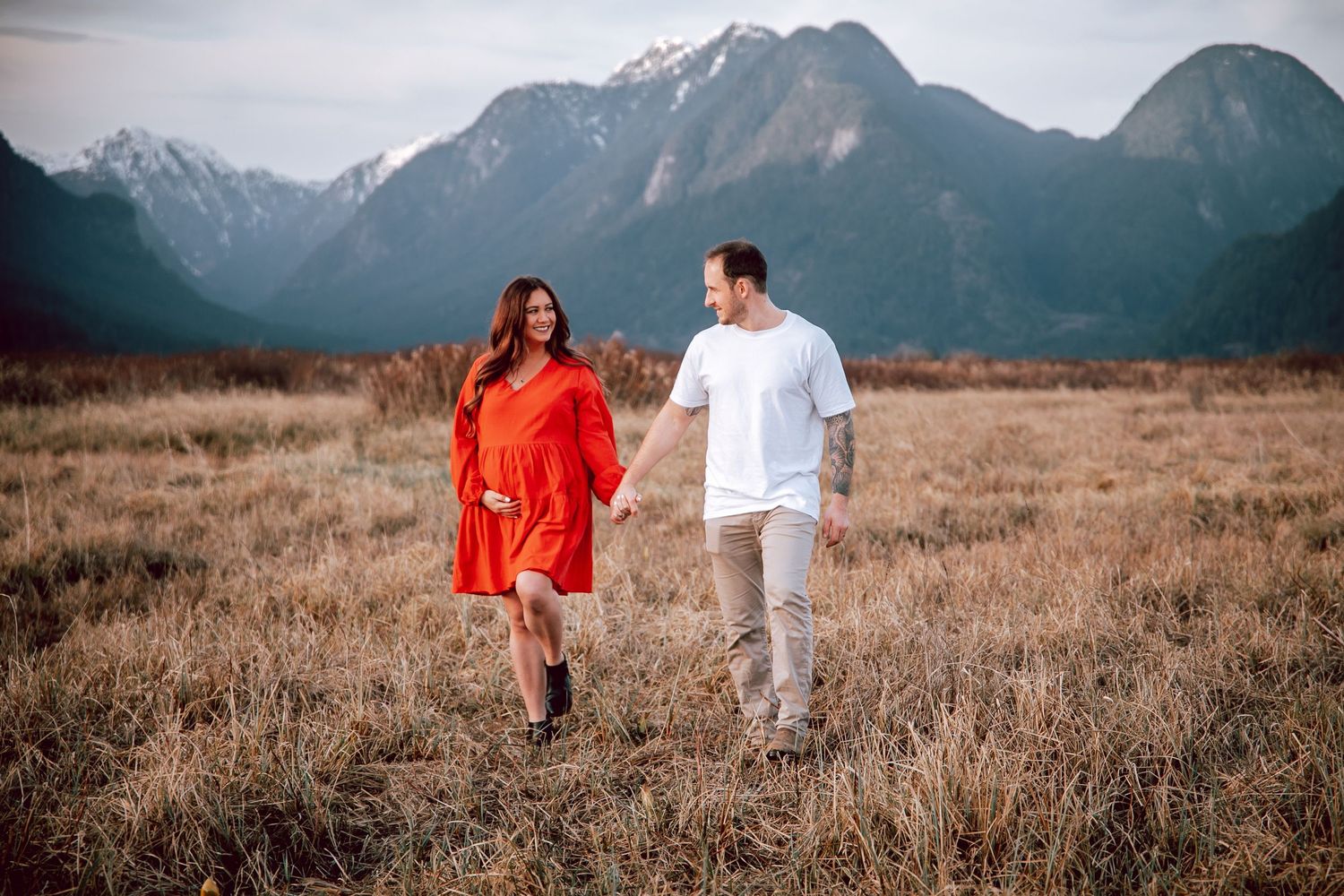 Couple in red dress and white outfit walking hand in hand through mountain valley field.