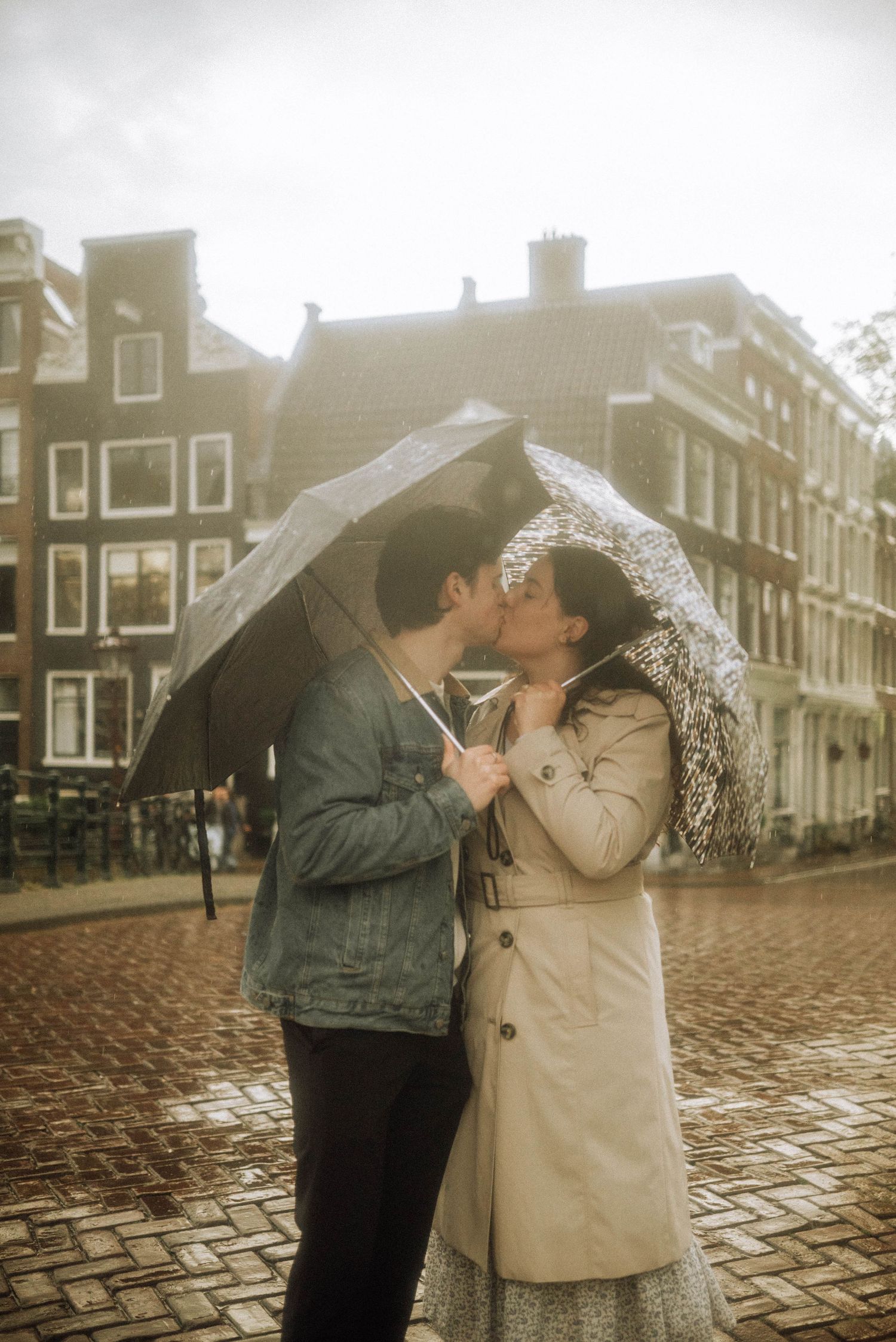 Couple kissing under a clear umbrella on a rainy cobblestone street - Brouwersgracht, Amsterdam.