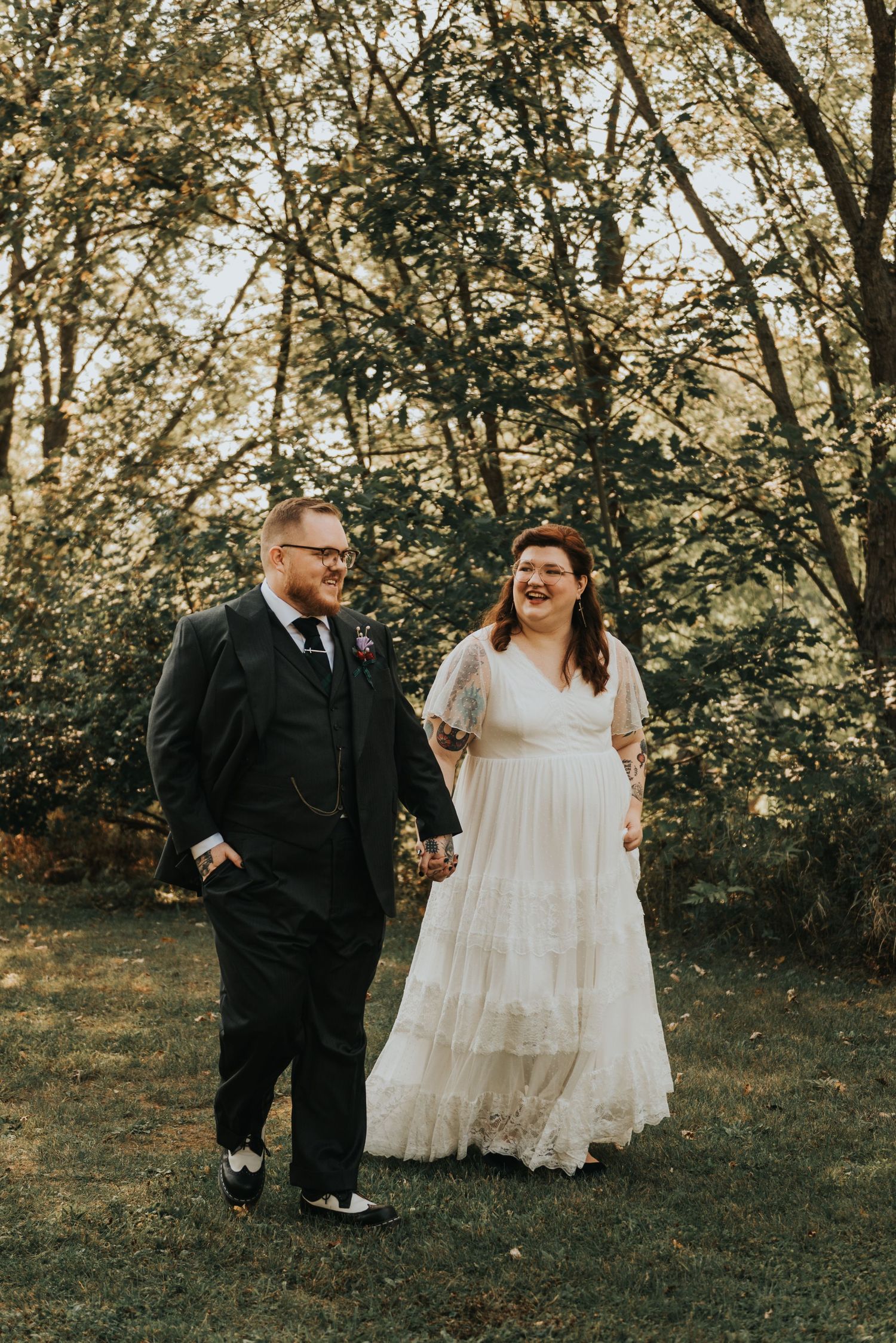 A wedding couple walks together and shares a joyful laugh while strolling through a sunlit wooded area.
