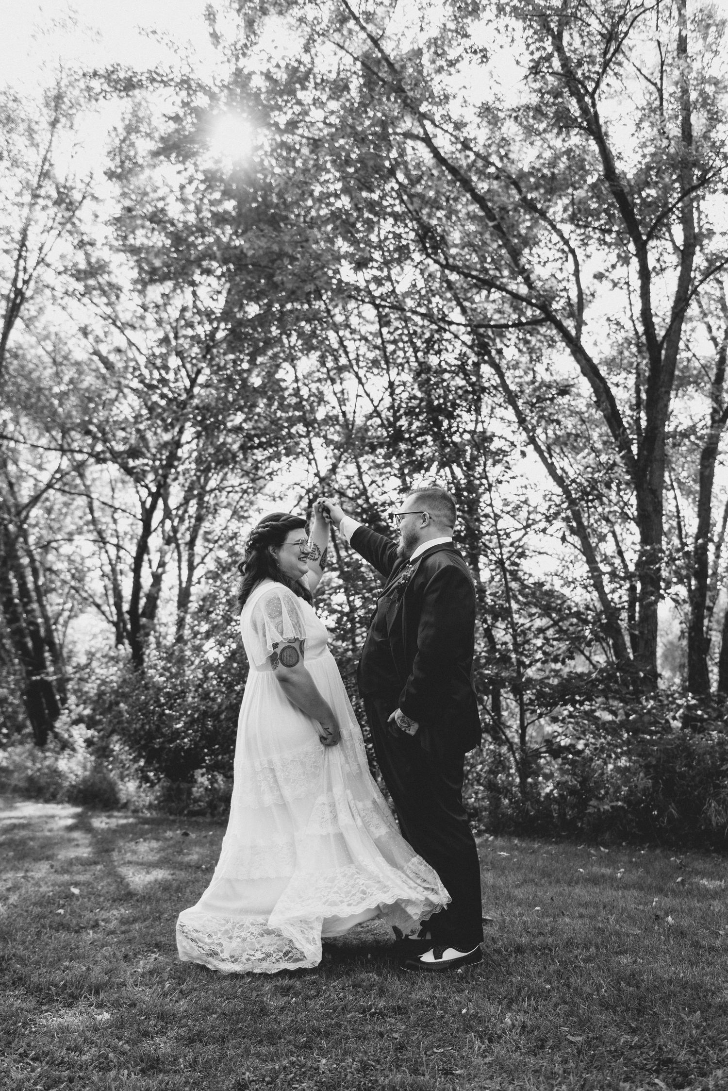 A romantic black and white wedding photograph showing a couple in formal attire standing together under sunlit trees.